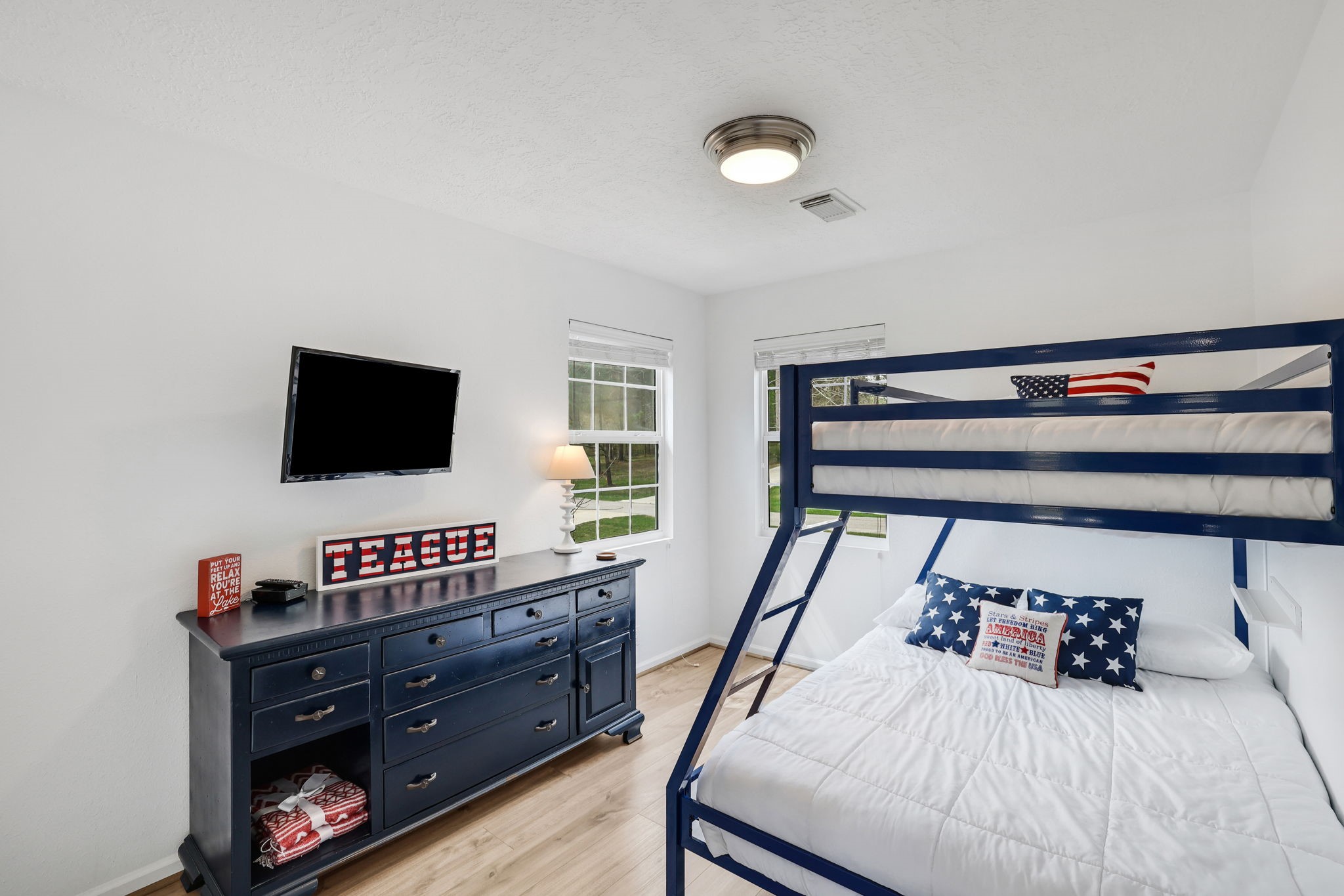 331 Holiday Lane Coldspring, TX 77331 - Photo 30 of 36 Bright secondary bedroom featuring a bunk bed with patriotic-themed bedding, a mounted TV, and a dark blue dresser. Natural light streams in through two windows, enhancing the room's cozy ambiance.