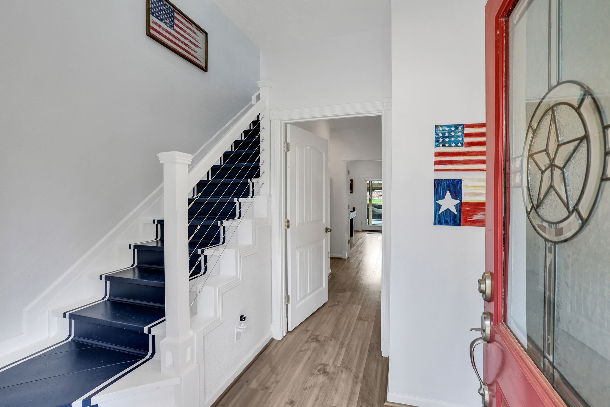 331 Holiday Lane Coldspring, TX 77331 - Photo 7 of 36 This inviting entryway features a red door with decorative glass, a staircase with navy and white accents, and wood flooring leading to a bright living area. American-themed decor adds a charming touch.