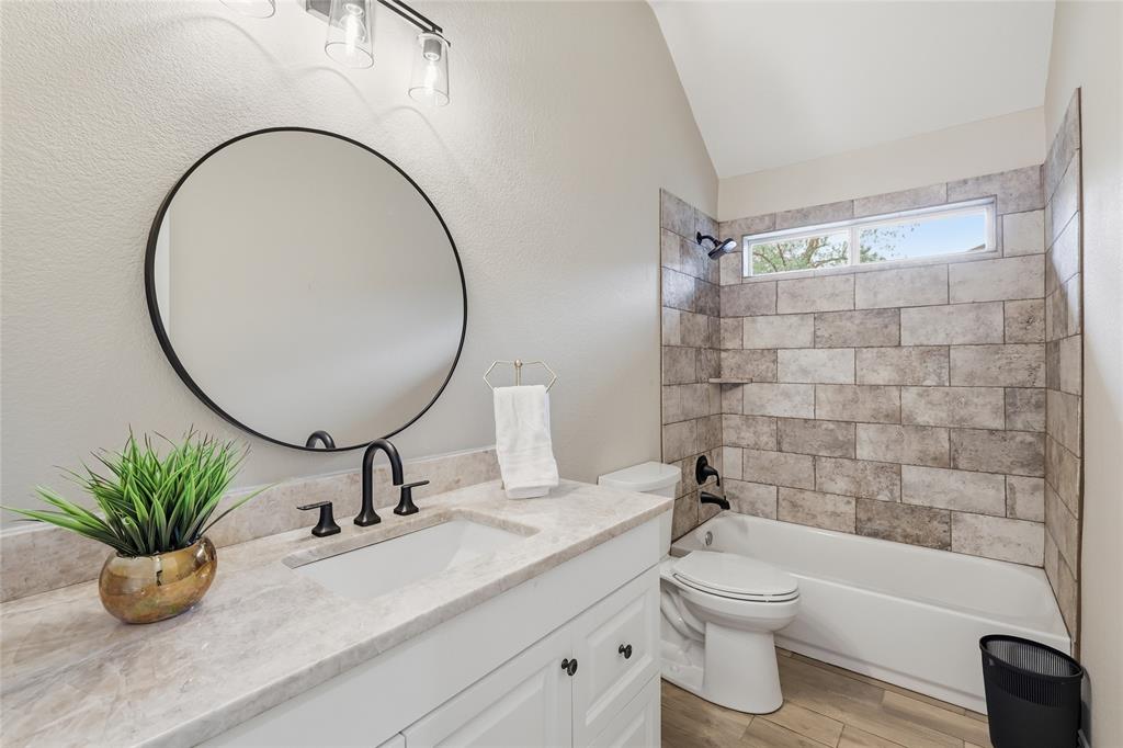 2402 Ravinia Drive Corinth, TX 76208 - Photo 31 of 40 Well-appointed bathroom featuring a vanity with a light-colored countertop and a circular mirror, a bathtub with tiled surround, and wood-look flooring