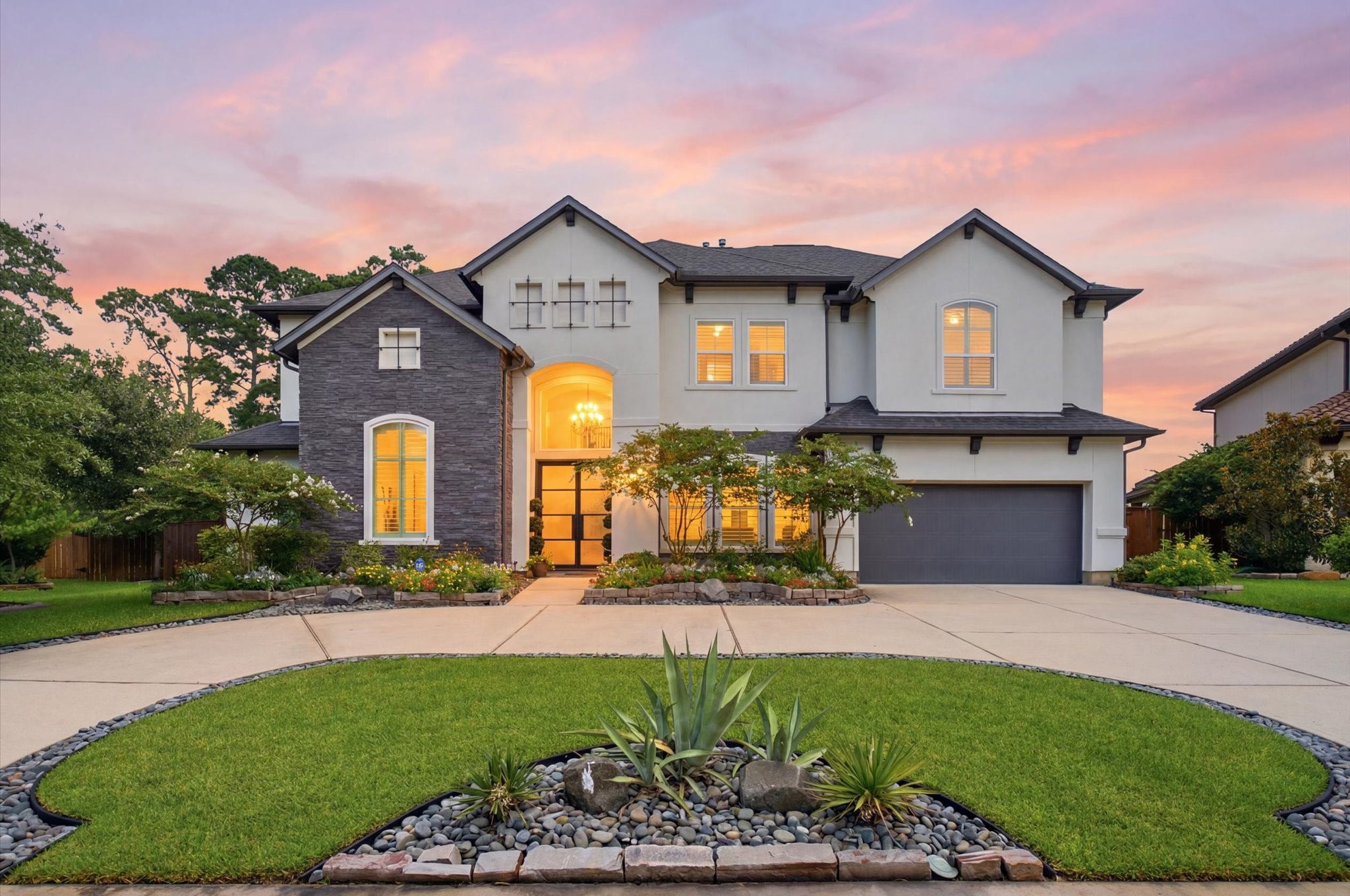 23722 Thortons Park Lane Spring, TX 77389 - Photo 1 of 48 This stunning two-story home features a modern exterior with a mix of stone and stucco finishes. It boasts large, illuminated windows, a spacious driveway leading to a two-car and a tandem garage, and beautifully landscaped front yard with vibrant greenery. The sunset backdrop adds a warm and inviting touch.