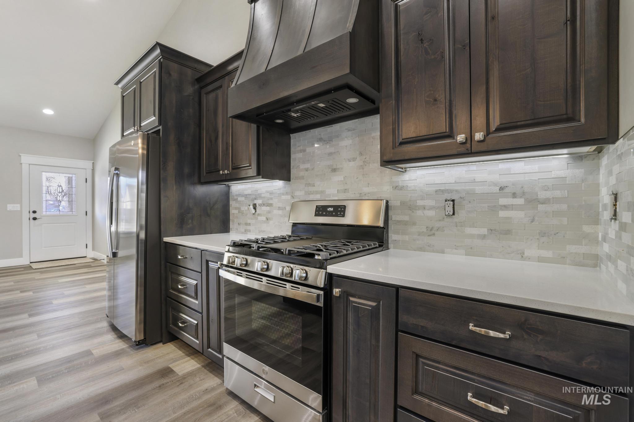 298 Sandi Road Twin Falls, ID 83301 - Photo 6 of 24 Modern kitchen featuring dark wood cabinetry, stainless steel appliances, a range hood, light-toned countertops, and a subway tile backsplash