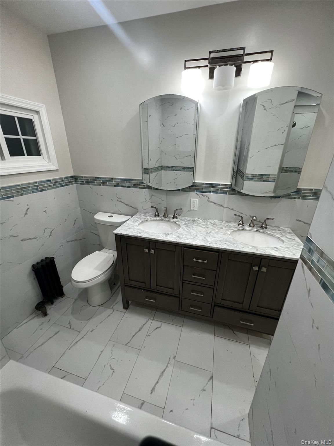 Bathroom featuring wainscoting, tile walls, double vanity, light marble finish flooring, and a tub