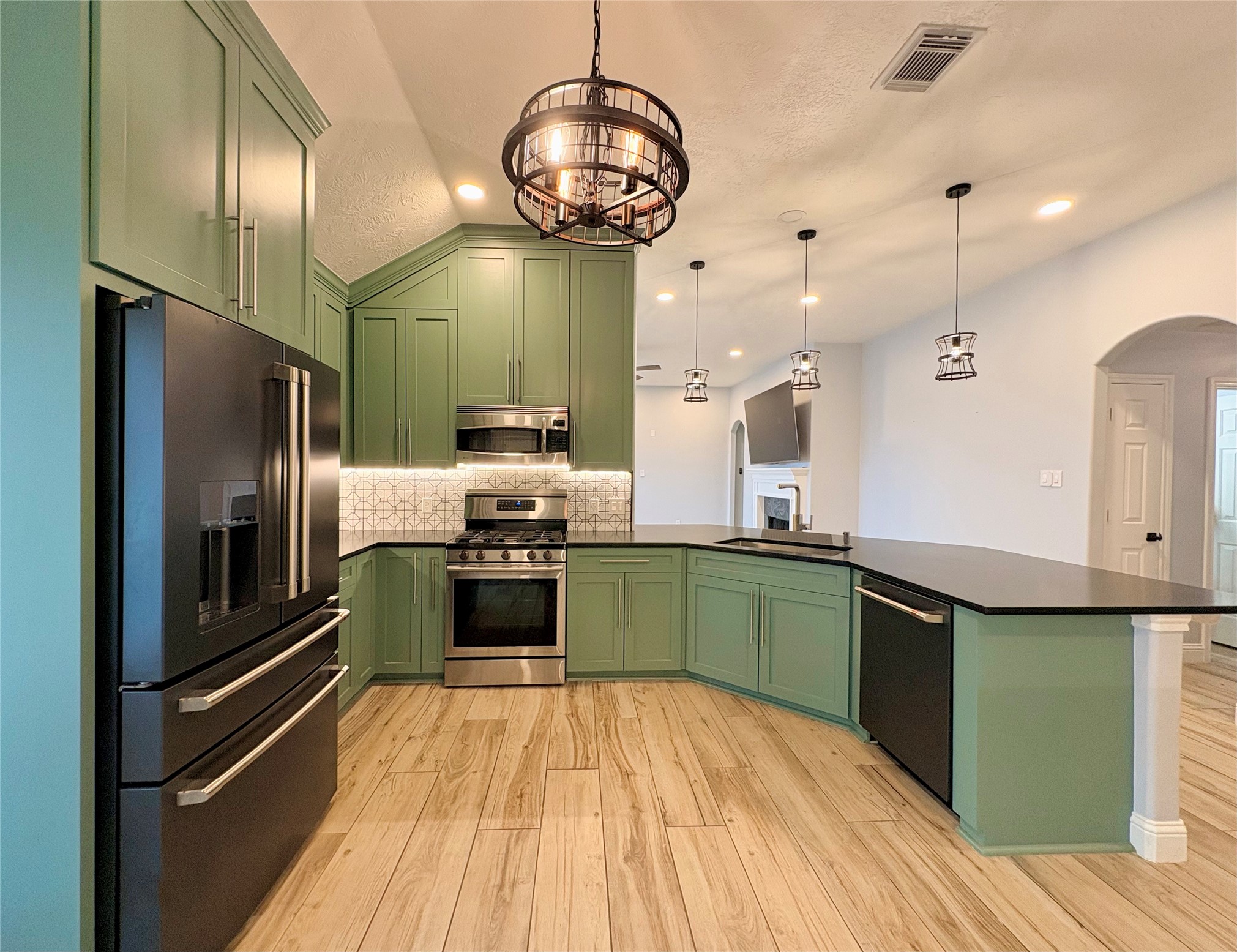 15410 Redbud Berry Way Cypress, TX 77433 - Photo 10 of 49 This kitchen features modern green cabinetry, stainless steel appliances, and a sleek black countertop. The open layout includes a large island with pendant lighting and wood-look flooring, creating a stylish and functional space.