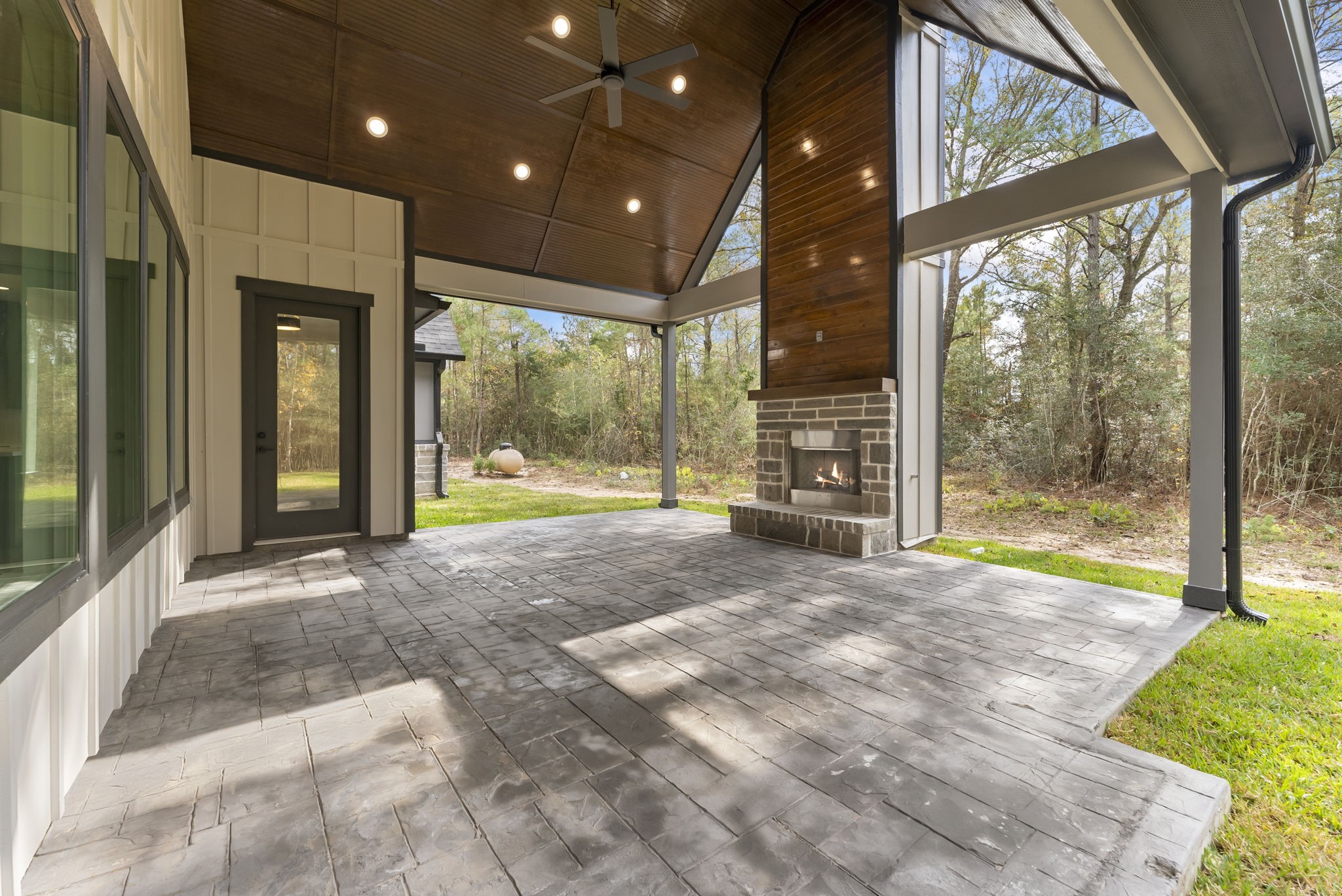 29001 Colin Court Waller, TX 77484 - Photo 36 of 47 This spacious covered patio features a high vaulted ceiling with recessed lighting and a ceiling fan, creating an inviting outdoor space. It includes a modern fireplace and offers a serene view of the surrounding wooded area, perfect for relaxation or entertaining.
