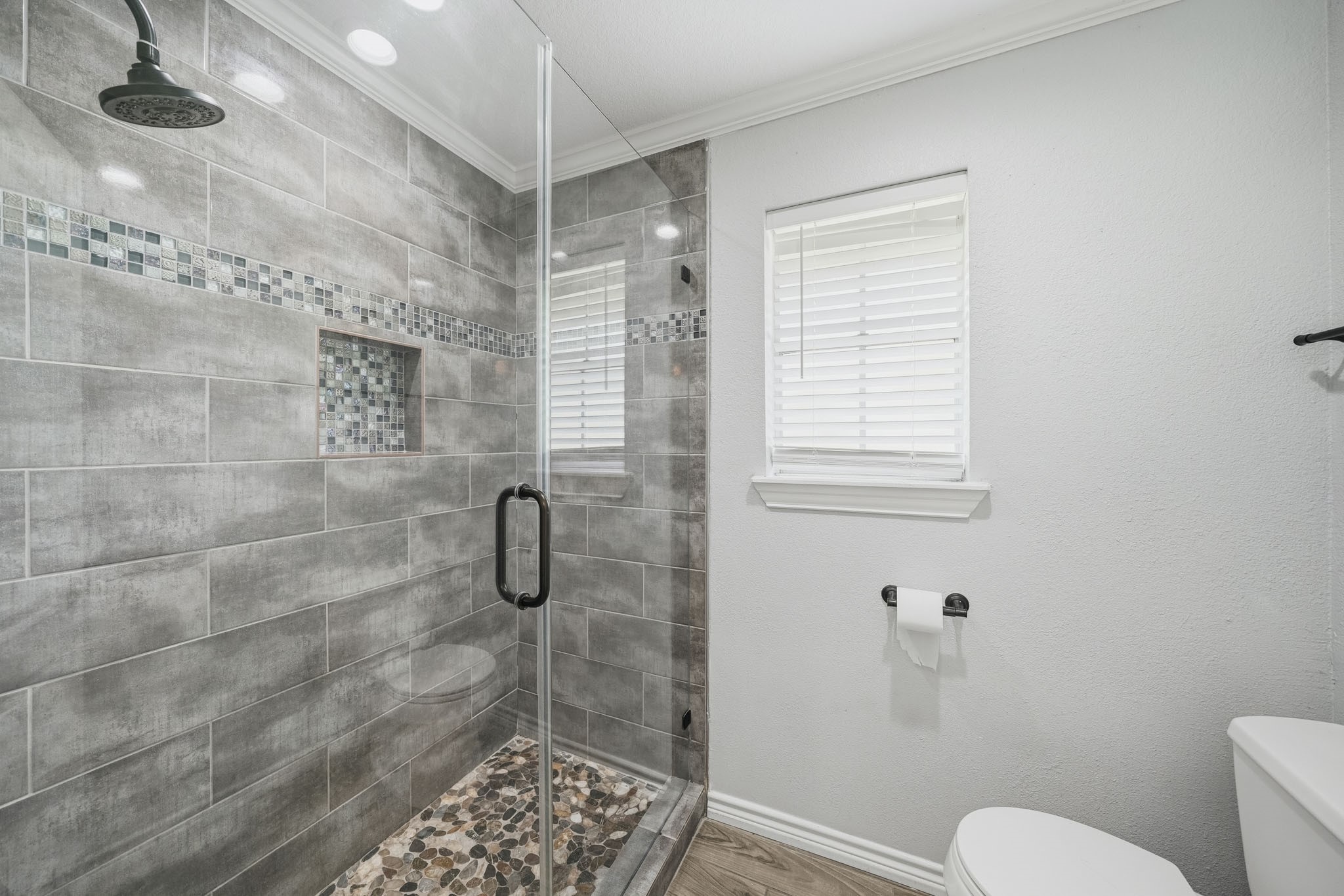 302 Country Road 893 Angleton, TX 77515 - Photo 22 of 26 This bathroom features a modern walk-in shower with gray tile and mosaic accents, a pebble stone floor, and a glass door. It has a window with blinds for natural light and a toilet nearby, creating a clean and elegant space.