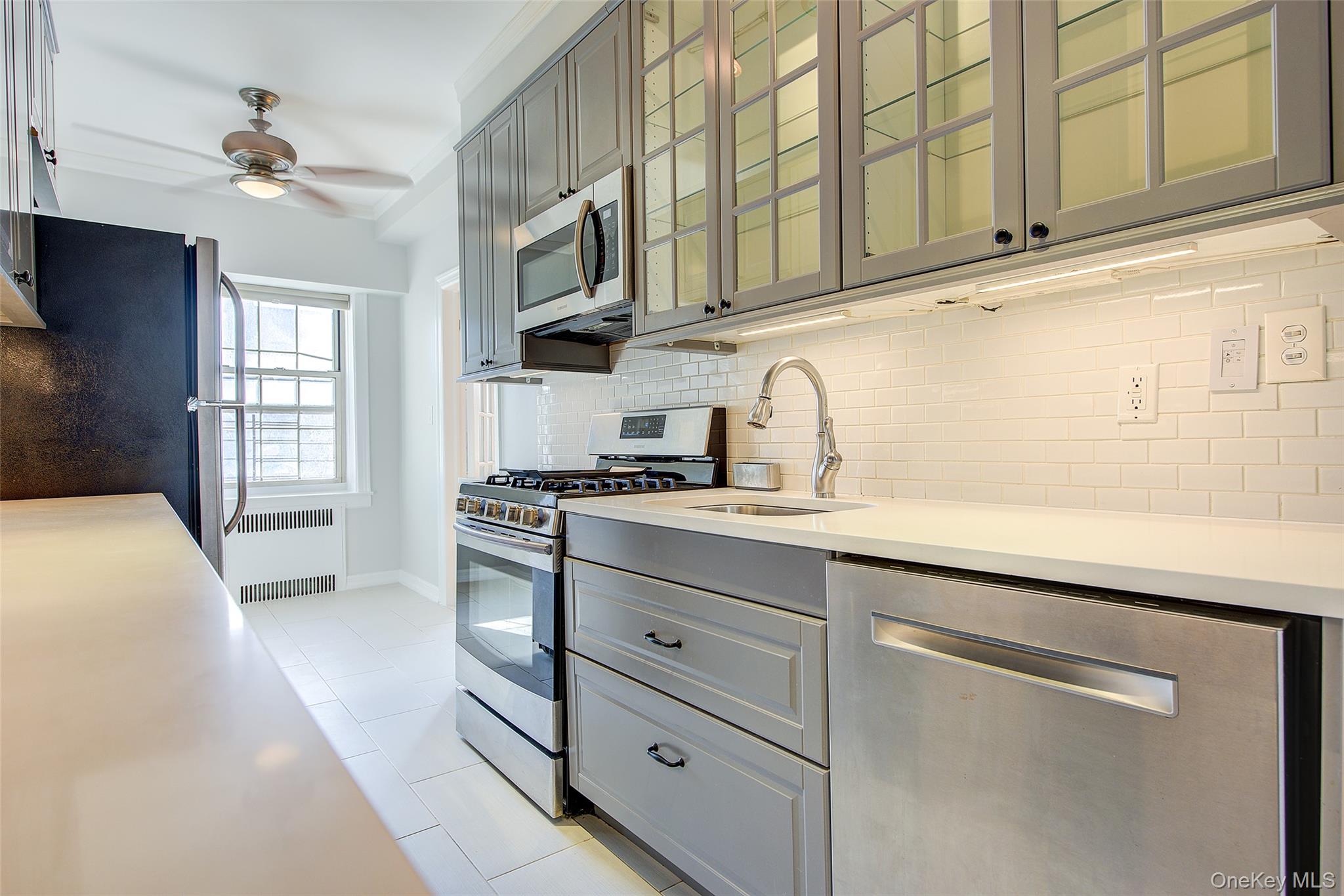 79-15 35th Avenue, Unit 5A Queens, NY 11372 - Photo 8 of 14 Kitchen with stainless steel appliances, gray cabinetry, glass insert cabinets, radiator, and backsplash