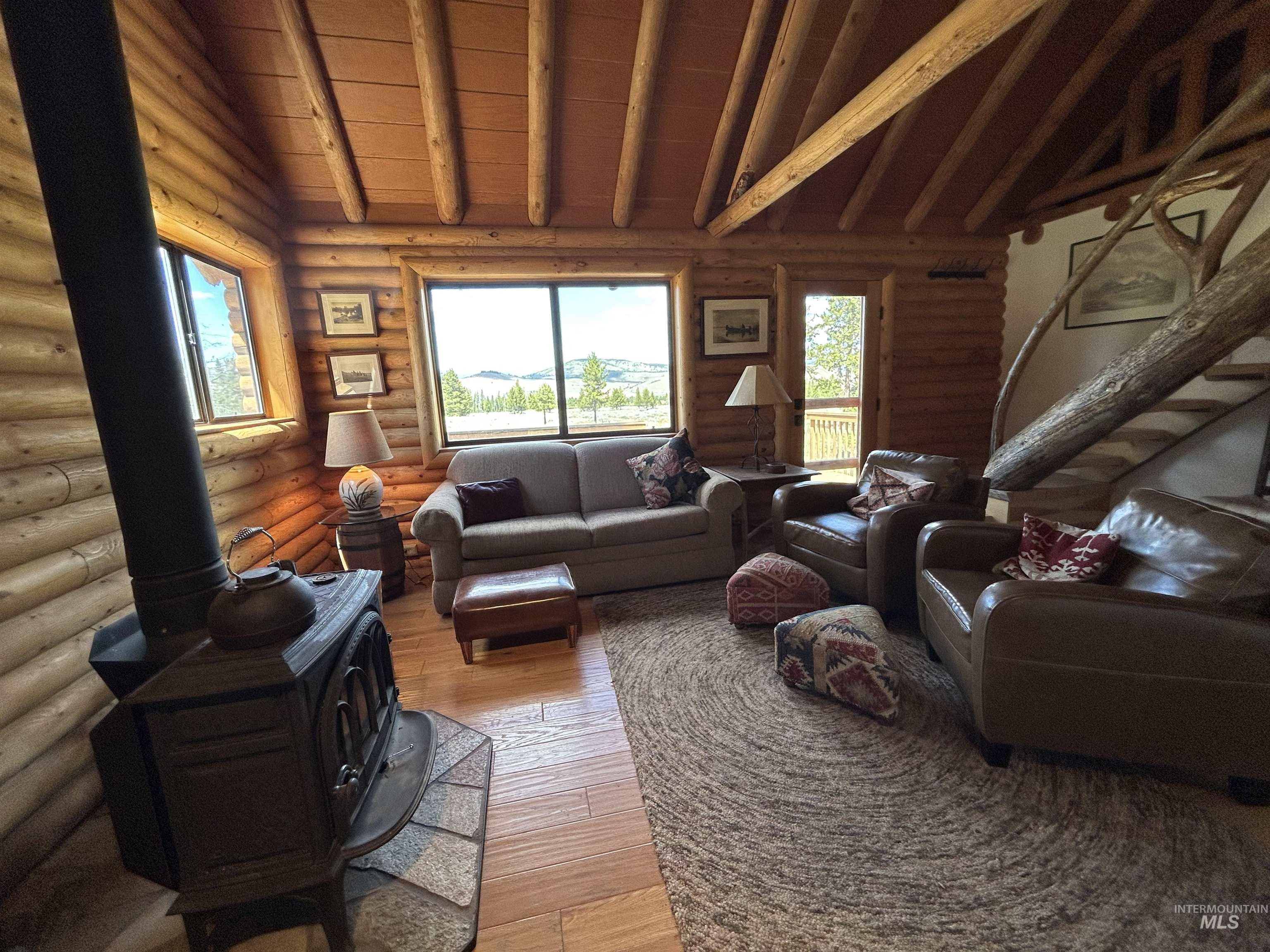 35 Sawtooth Way Stanley, ID 83278 - Photo 14 of 36 Living area featuring log wall construction, exposed beam ceiling, large windows, a freestanding wood-burning stove with a stone hearth, and a log staircase with wooden steps