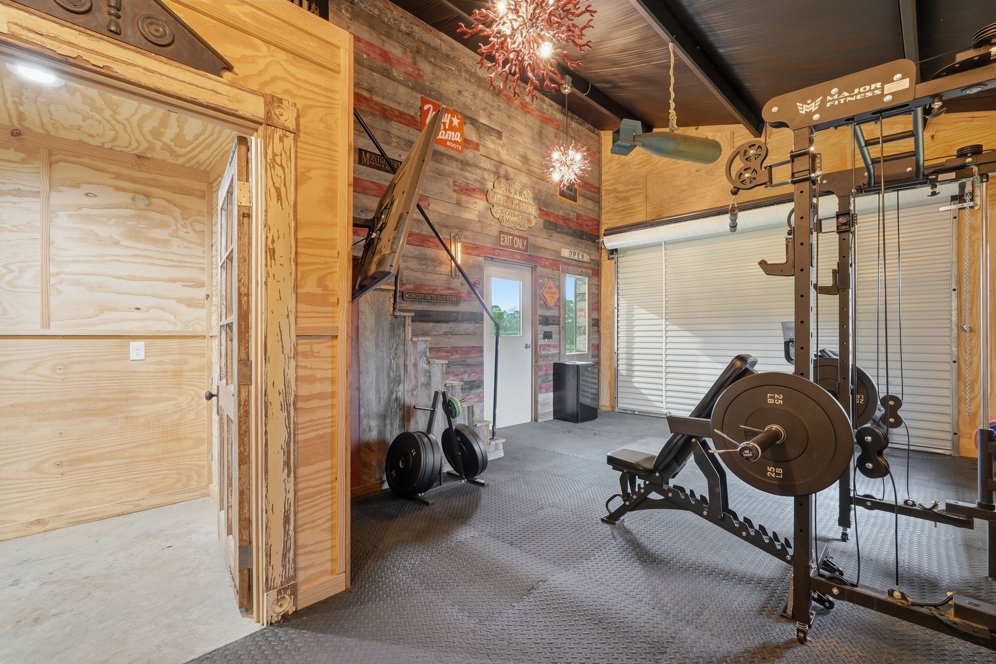 2743 Bobville Road Montgomery, TX 77316 - Photo 49 of 50 This photo showcases a home gym with exercise equipment on rubber flooring, wood-paneled walls, and a rustic aesthetic. It features a large garage door for natural light and ventilation, creating an ideal workout space.