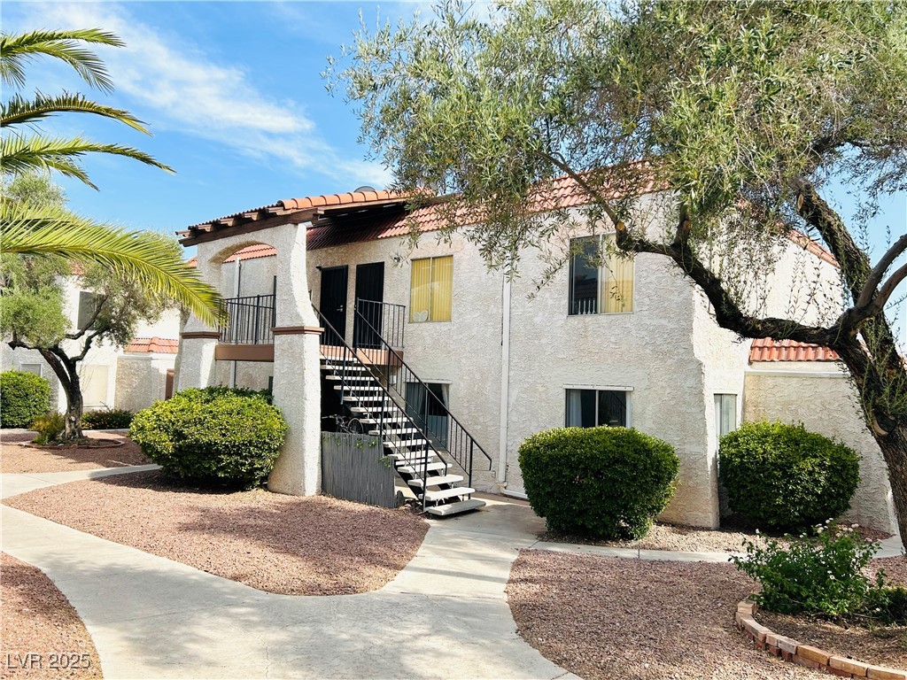 524 Fir Street, Unit 203 Boulder City, NV 89005 - Photo 7 of 7 This charming ground floor condo features a red-tiled roof and stucco walls, complemented by a well-maintained front yard with mature olive trees and neatly trimmed bushes. A concrete walkway with black metal railings leads to the inviting entrance, enhanced by a bright, sunny day.