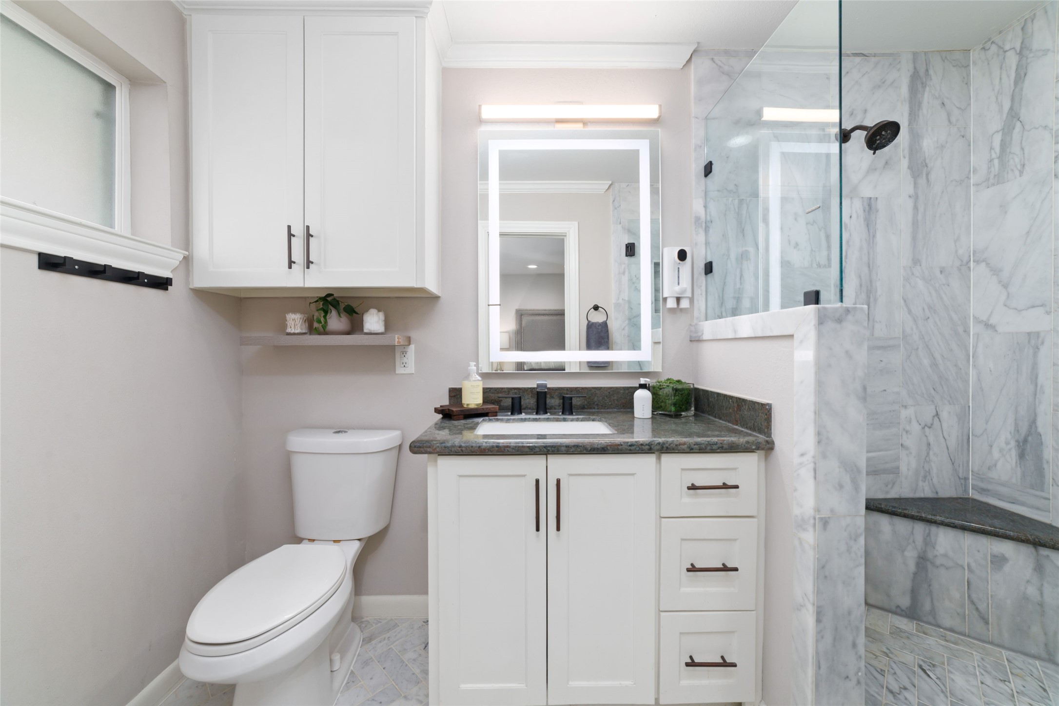 10607 Chimney Rock Road Houston, TX 77096 - Photo 28 of 35 This wasn’t left as a standard bath… it was opened up and reworked into something that actually feels like a primary suite should. Oversized walk-in marble shower with dual heads, built-in seat, and a full glass wall that keeps it open and clean. LED mirror, added cabinetry for real storage, natural light from the window, and marble herringbone floors that pull it all together… it’s finished in a way you feel every day.