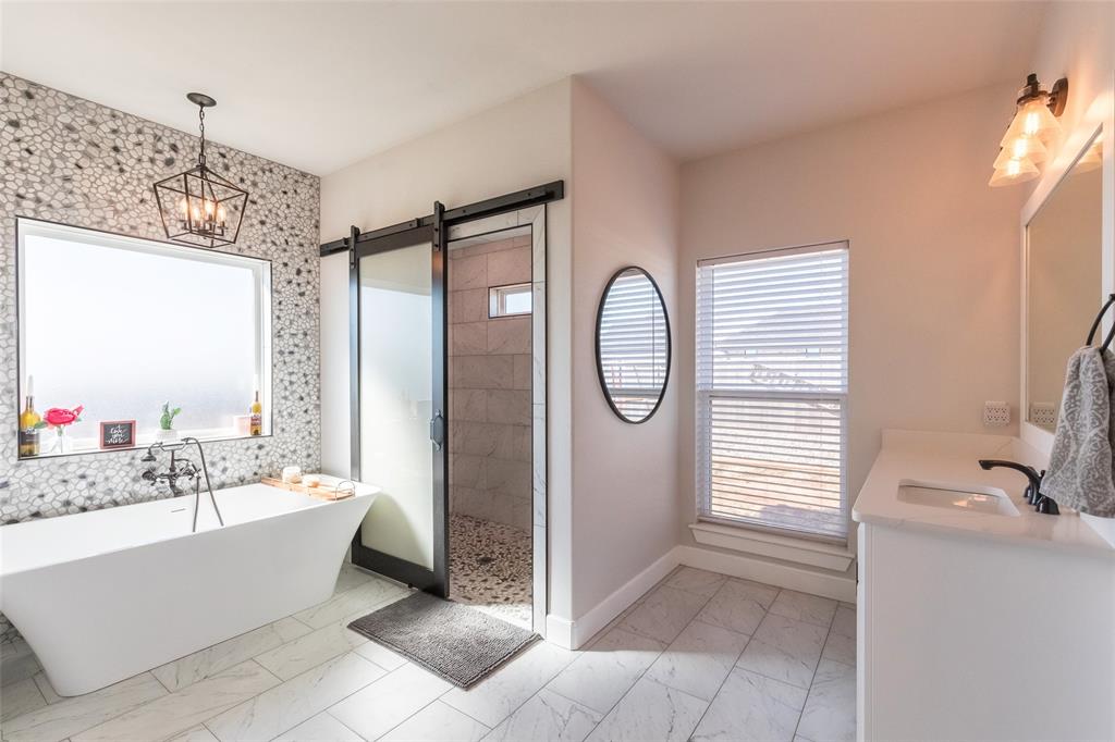 107 Kleingrass Road Abilene, TX 79606 - Photo 13 of 33 Freestanding soaking tub with a wall-mounted faucet, pebble-tile accent wall, clear glass barn door shower enclosure, vanity with integrated sink, and tile flooring