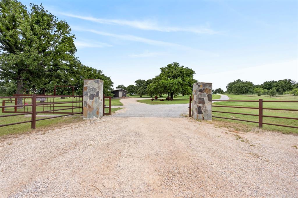 1080 North Greenbrier Road Nocona, TX 76255 - Photo 40 of 40 Gravel driveway entry framed by stone pillars and a metal gate, opening to a sprawling property with mature trees and a distant outbuilding