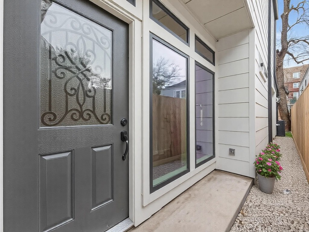 1319 West Bell Street Houston, TX 77019 - Photo 5 of 50 This photo showcases a modern entryway with a decorative glass front door and large windows, allowing natural light to enhance the space. The exterior features light siding and a small gravel walkway lined with a wooden fence and a potted plant, providing a welcoming and stylish entrance.