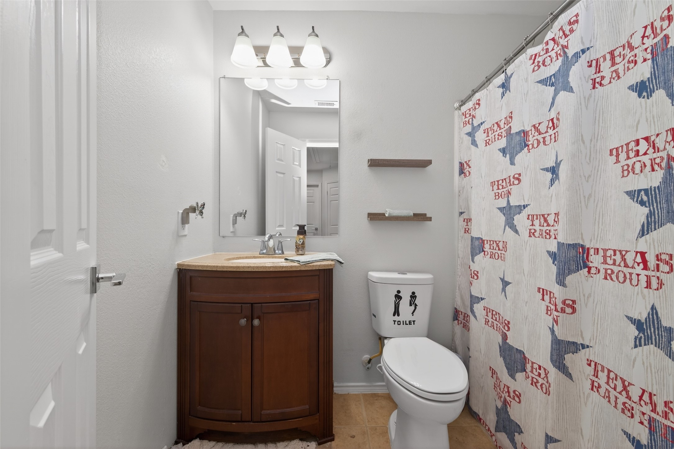 20218 Appaloosa Ridge Drive Humble, TX 77338 - Photo 24 of 44 This bathroom features a cozy design with a wooden vanity, large mirror, and overhead lighting. The shower curtain has a Texas theme, and there are minimalist shelves above the toilet for additional storage.