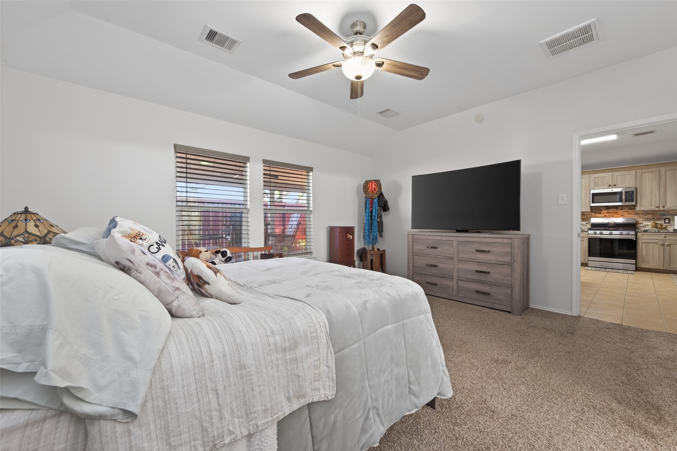 20218 Appaloosa Ridge Drive Humble, TX 77338 - Photo 28 of 44 This bedroom is spacious and bright, featuring neutral carpeting and walls, a ceiling fan, and large windows with wooden blinds. It includes a comfortable bed, a large dresser with a TV, and an adjoining view into a modern kitchen.