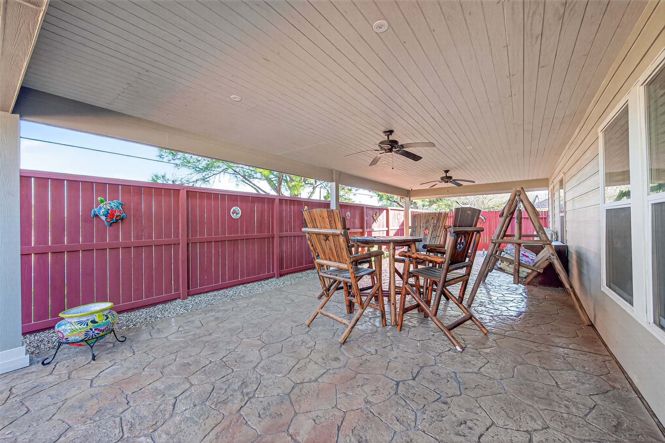 20218 Appaloosa Ridge Drive Humble, TX 77338 - Photo 35 of 44 This photo showcases a spacious covered patio with a textured stone floor, wooden ceiling, and ceiling fans. It features a high-top table with chairs and a wooden swing, perfect for outdoor relaxation and entertaining. The area is enclosed with a red wooden fence, offering privacy.
