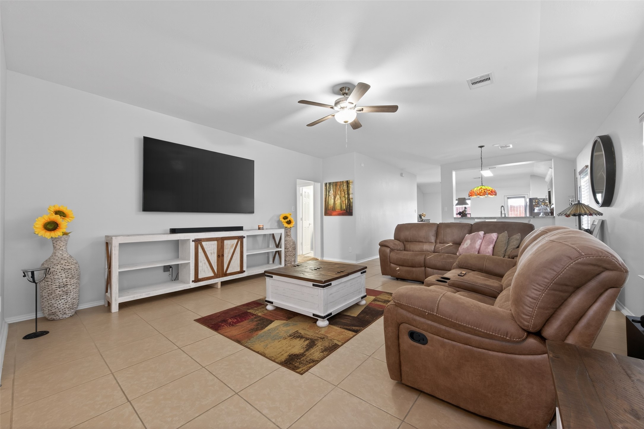 20218 Appaloosa Ridge Drive Humble, TX 77338 - Photo 10 of 44 This spacious living room features tiled floors, a large wall-mounted TV, and comfortable brown recliner seating. The space is bright and inviting with neutral walls, ceiling fan, and decorative touches like sunflowers and a colorful area rug. There's a view into the open kitchen, enhancing the home's open-concept feel.