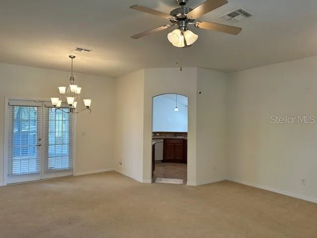 an empty room with chandelier fan and kitchen view