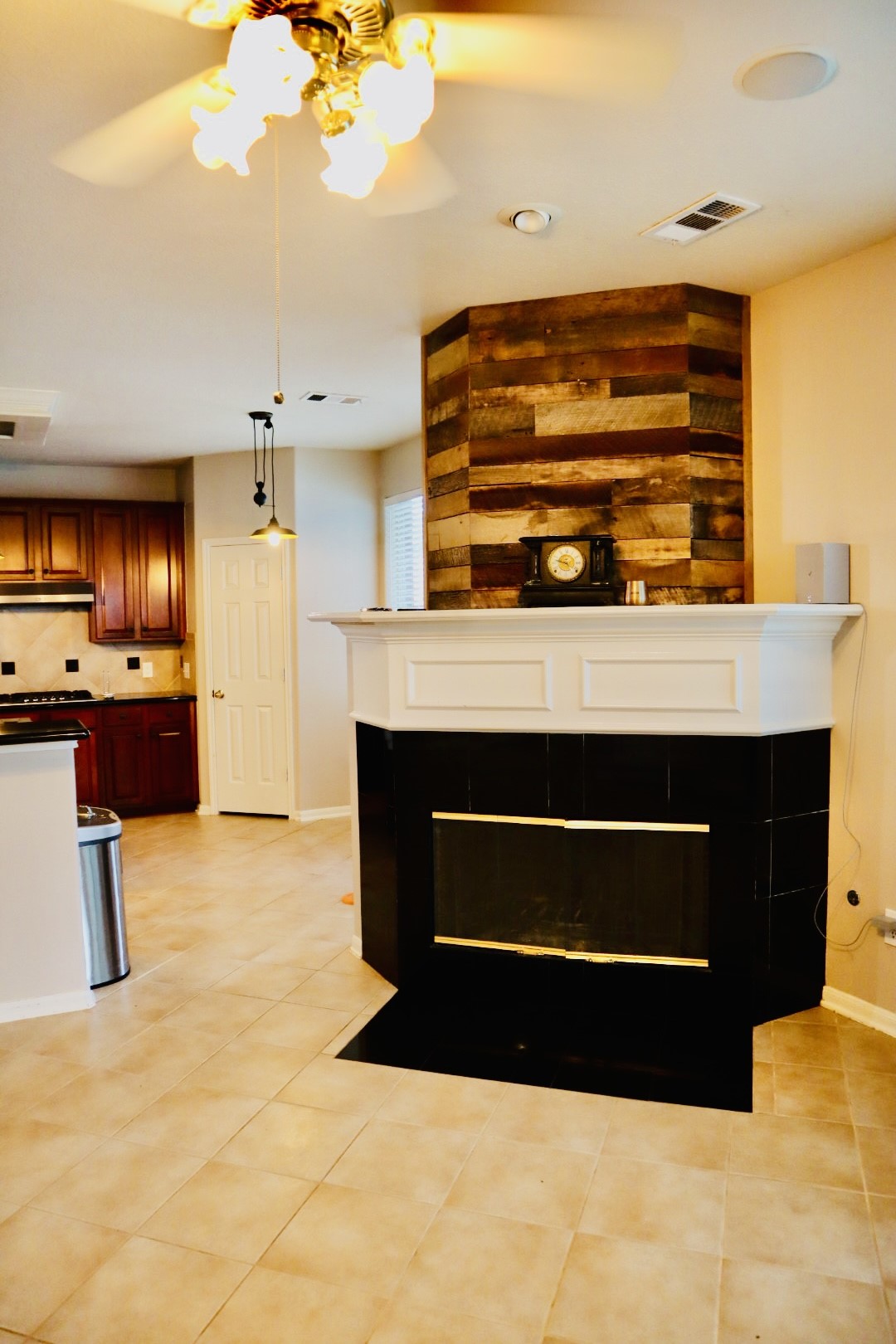 19114 Emory Trail Spring, TX 77388 - Photo 11 of 32 Family Room shows a cozy living space with a modern corner fireplace featuring a wood accent wall. The room has tiled flooring, warm lighting, and opens into a kitchen area with wooden cabinets, offering a welcoming and functional layout.