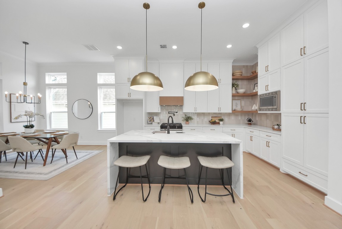 This modern kitchen features elegant white cabinetry, a large marble island with seating, and stylish pendant lighting. The open layout flows into a dining area with ample natural light, creating a bright and inviting space.