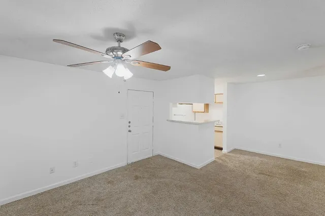 an empty room with chandelier fan and kitchen view