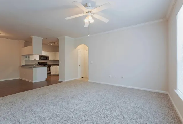 an empty room with chandelier fan and kitchen view