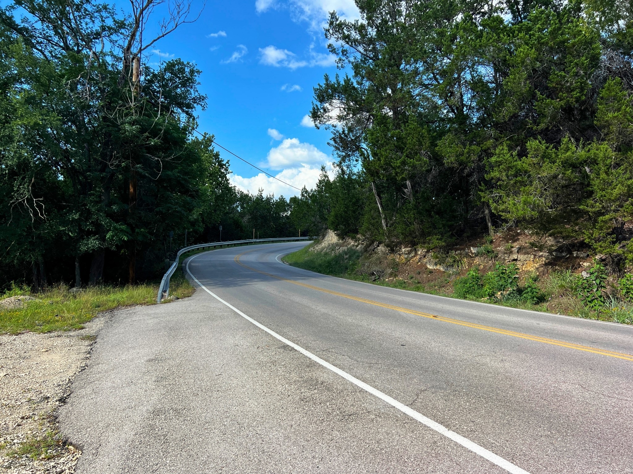 4500 Outpost Trace Leander, TX 78645 - Photo 12 of 13 A gently curving road winds through a lush, tree-lined landscape under a bright, cloud-dotted sky.