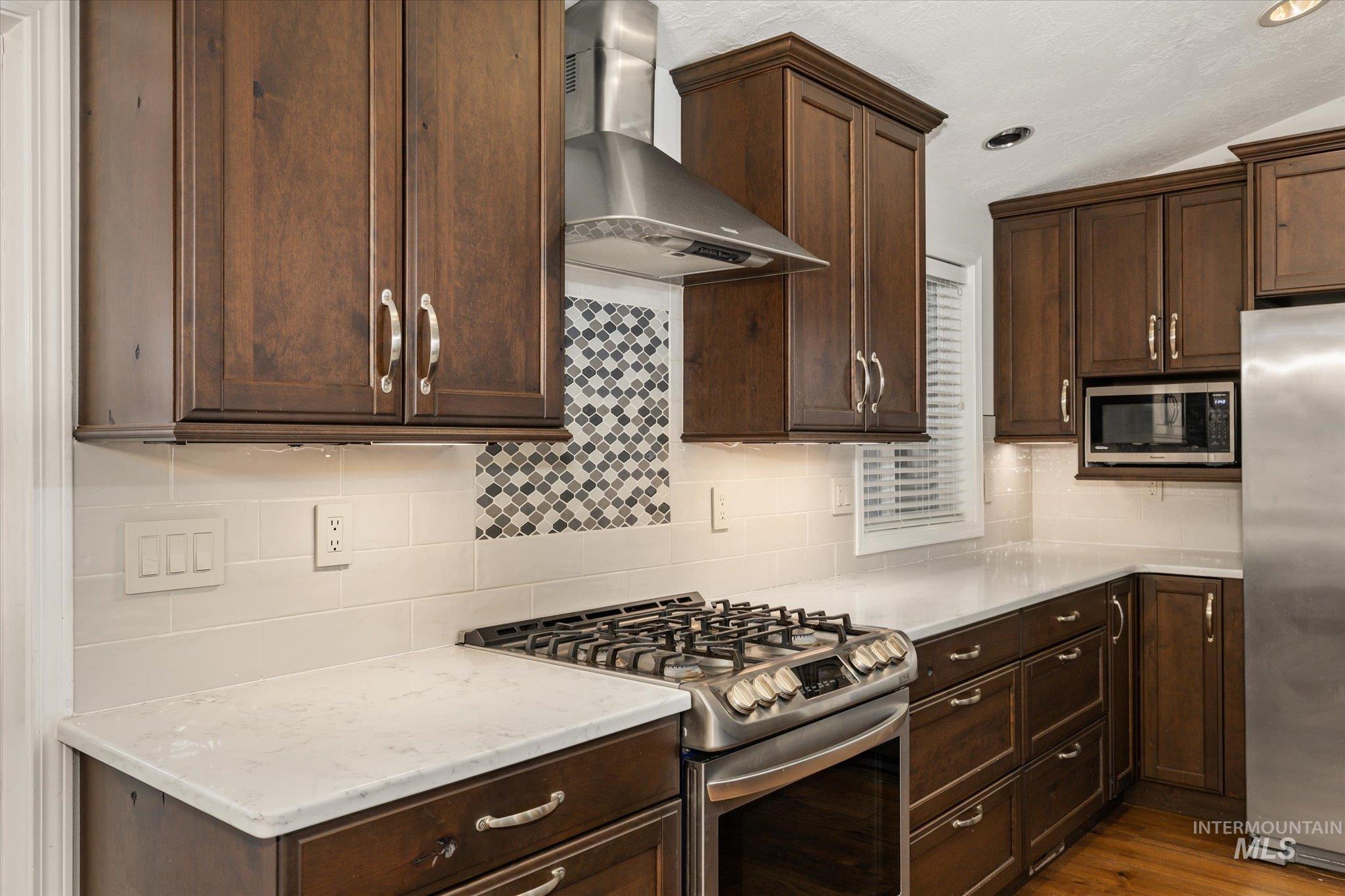 2870 North Morello Avenue Meridian, ID 83646 - Photo 19 of 20 Kitchen featuring appliances with stainless steel finishes, wall chimney range hood, dark brown cabinetry, backsplash, and light stone countertops