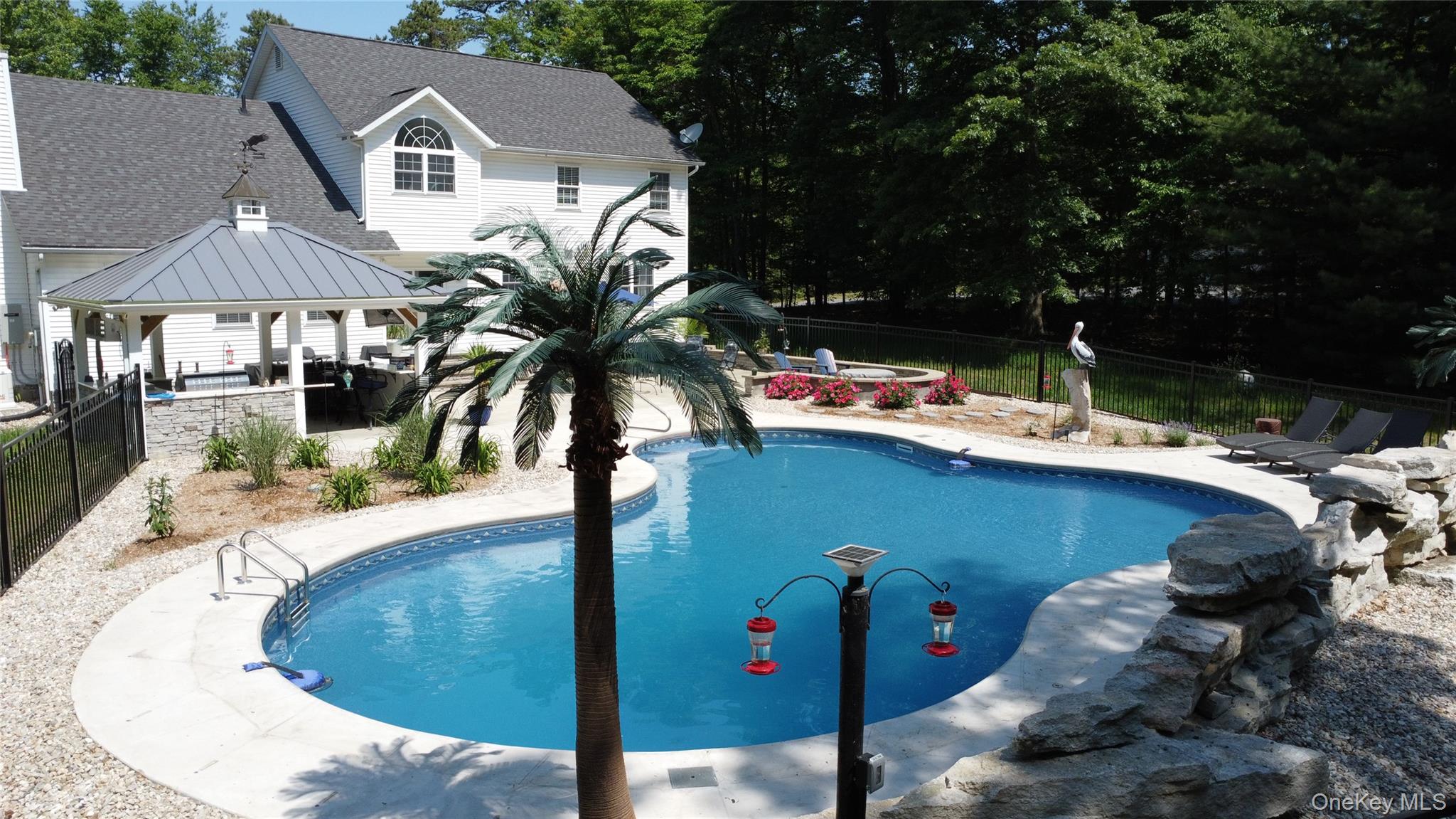 95 Weed Road Pine Bush, NY 12566 - Photo 25 of 31 Freeform salt water swimming pool featuring a white concrete surround, adjacent stone retaining wall, and a nearby covered patio structure with a dark roof