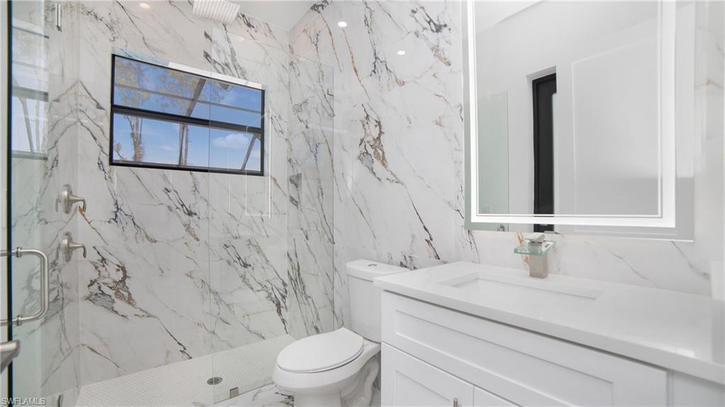 2343 Oil Well Road Naples, FL 34120 - Photo 22 of 46 Bathroom featuring full-height marble-style tiled walls, a glass-enclosed shower with a rainfall showerhead, a single vanity with a white countertop and base cabinetry, and an illuminated mirror