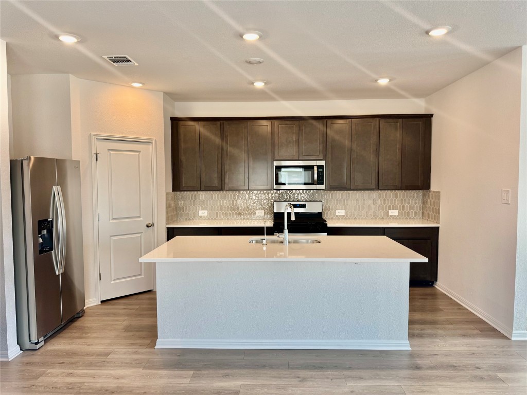 133 Boondoggle Ridge Cedar Creek, TX 78612 - Photo 2 of 19 Kitchen featuring dark brown cabinetry, appliances with stainless steel finishes, light stone counters, decorative backsplash, and an island with sink