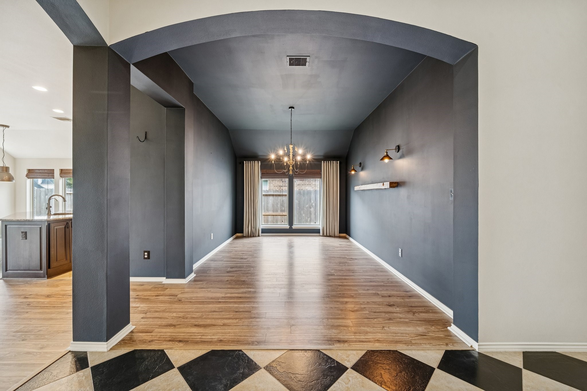 13014 Barkley Bend Lane Houston, TX 77044 - Photo 12 of 42 Defined by dramatic navy walls and a vaulted ceiling, this spacious formal dining room accommodates a large table for hosting guests. A wide arched entry and wood-inspired flooring create a seamless transition to the kitchen, while a classic chandelier and modern sconces provide elegant layers of light. Large windows offer a view of the outdoors, maintaining a bright atmosphere within this sophisticated space.