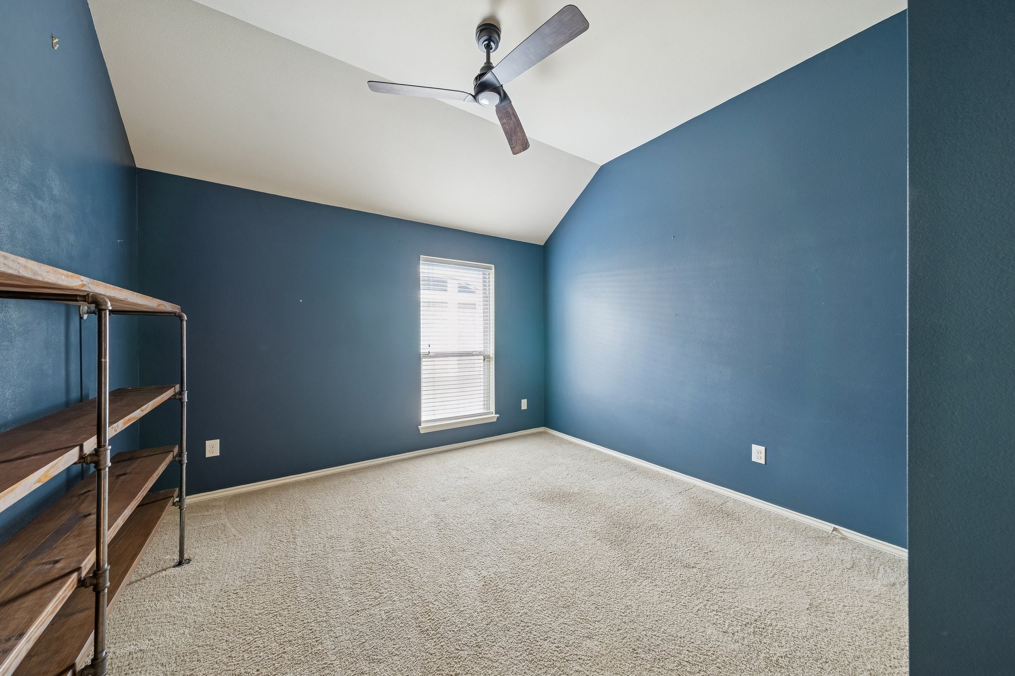 13014 Barkley Bend Lane Houston, TX 77044 - Photo 33 of 42 Deep navy blue walls and a vaulted ceiling create a sophisticated atmosphere in this versatile secondary bedroom or flex space. Plush neutral carpeting and a modern wood-blade ceiling fan ensure comfort, while the tall window provides a focused view of the outdoors. Industrial-inspired open shelving offers a unique storage solution that complements the room’s bold, contemporary aesthetic.