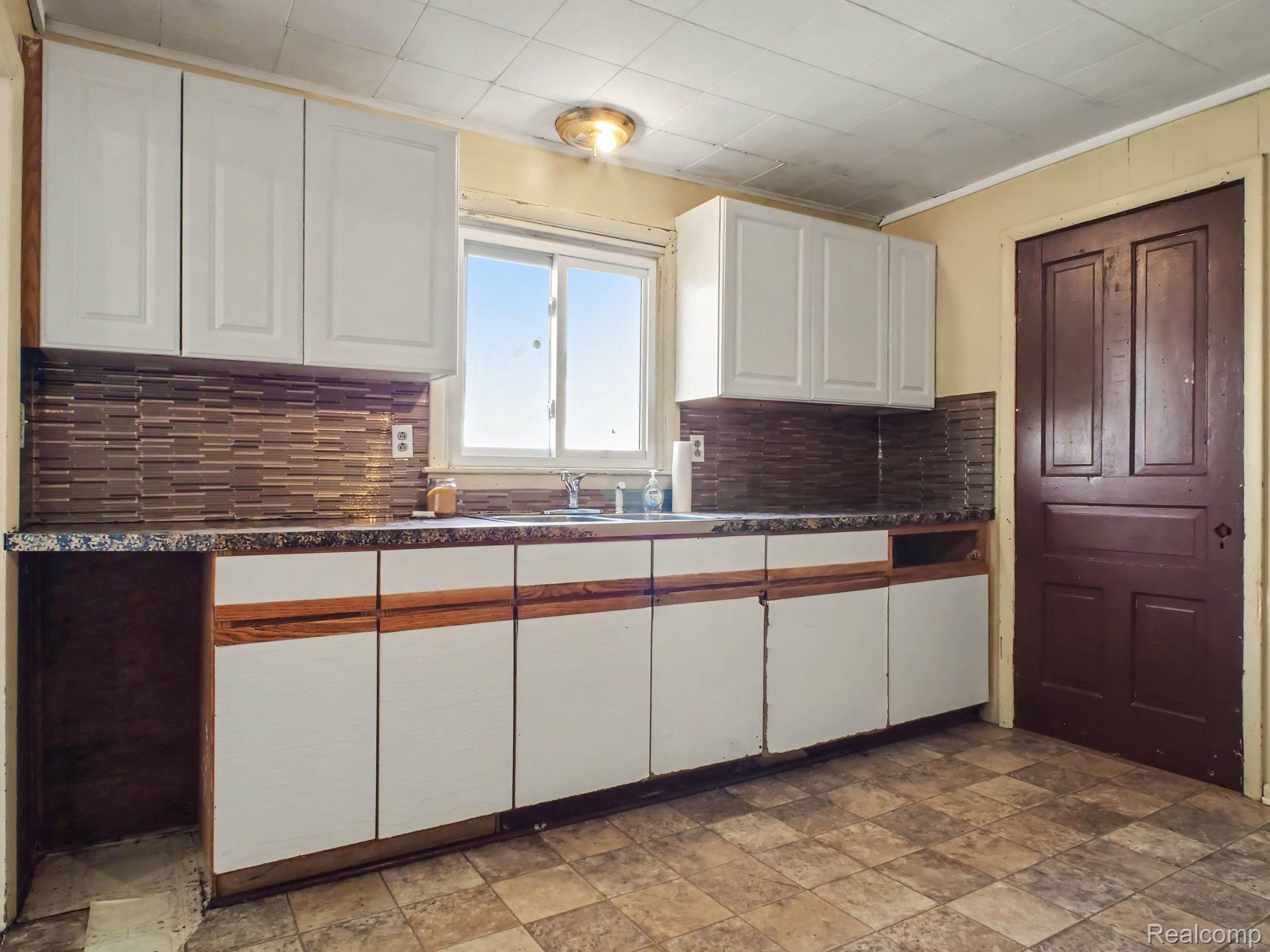 Spacious kitchen featuring white cabinetry with wood-tone base accents, dark tile backsplash, granite countertops, and a large window allowing natural light