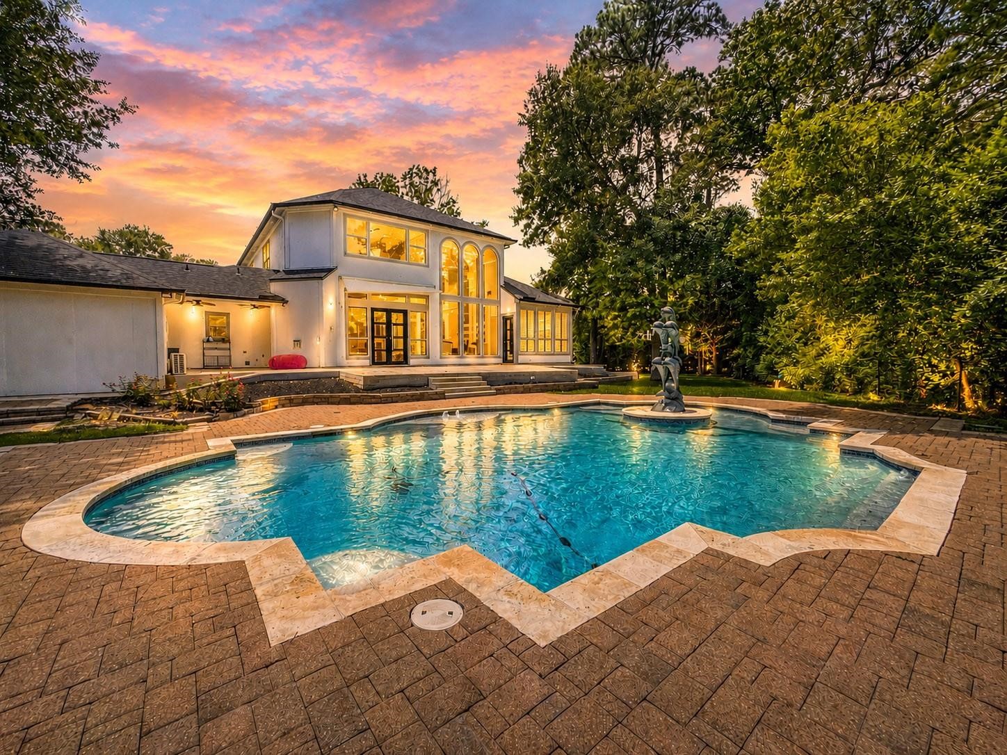 1810 Woerner Road Houston, TX 77090 - Photo 1 of 39 This photo showcases a luxurious two-story home with large windows and elegant architecture, set against a stunning sunset. The property features a beautifully landscaped backyard with a spacious, uniquely shaped swimming pool, surrounded by a paved patio and lush greenery, creating a serene and inviting outdoor space.