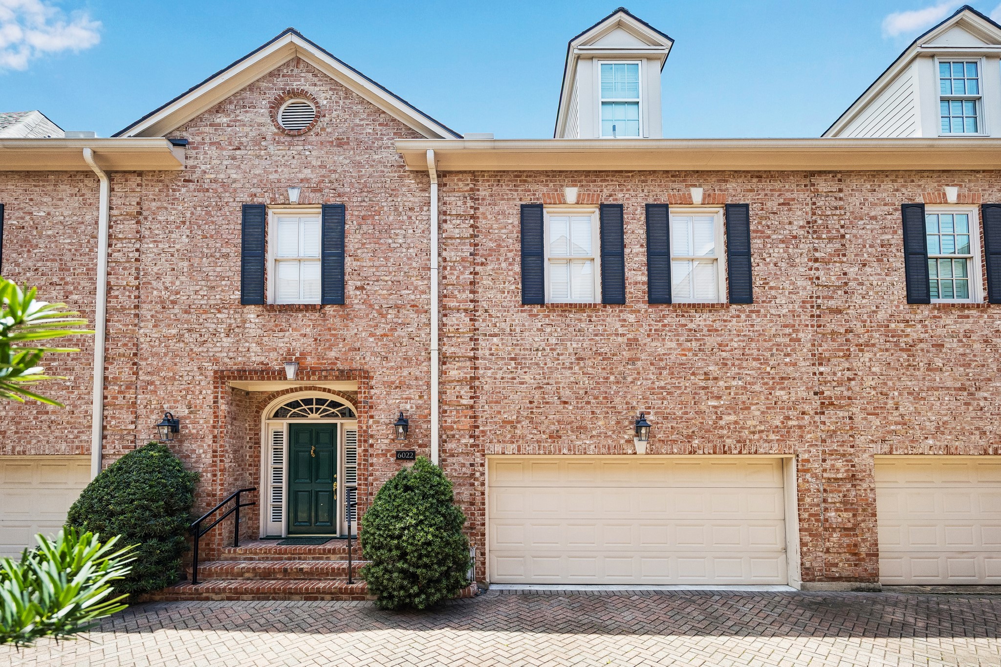 Classic brick townhome with timeless architecture, black shutters, and a welcoming entry framed by manicured landscaping and brick-paver drive.