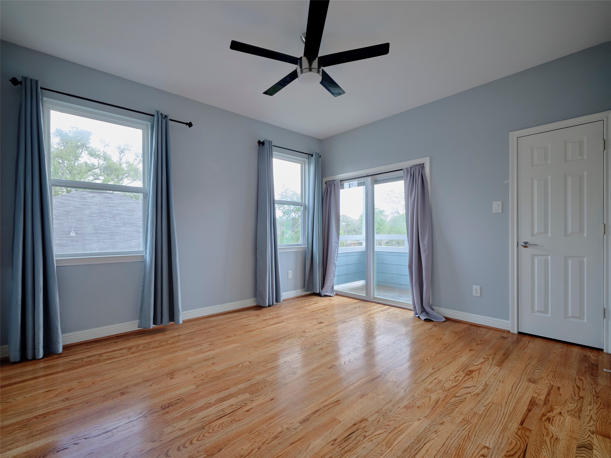 4219 Redbud Street Houston, TX 77051 - Photo 5 of 25 Bright, spacious room with hardwood floors, light blue walls, and large windows. It features a ceiling fan, sliding glass doors leading to a small balcony, and ample natural light.
