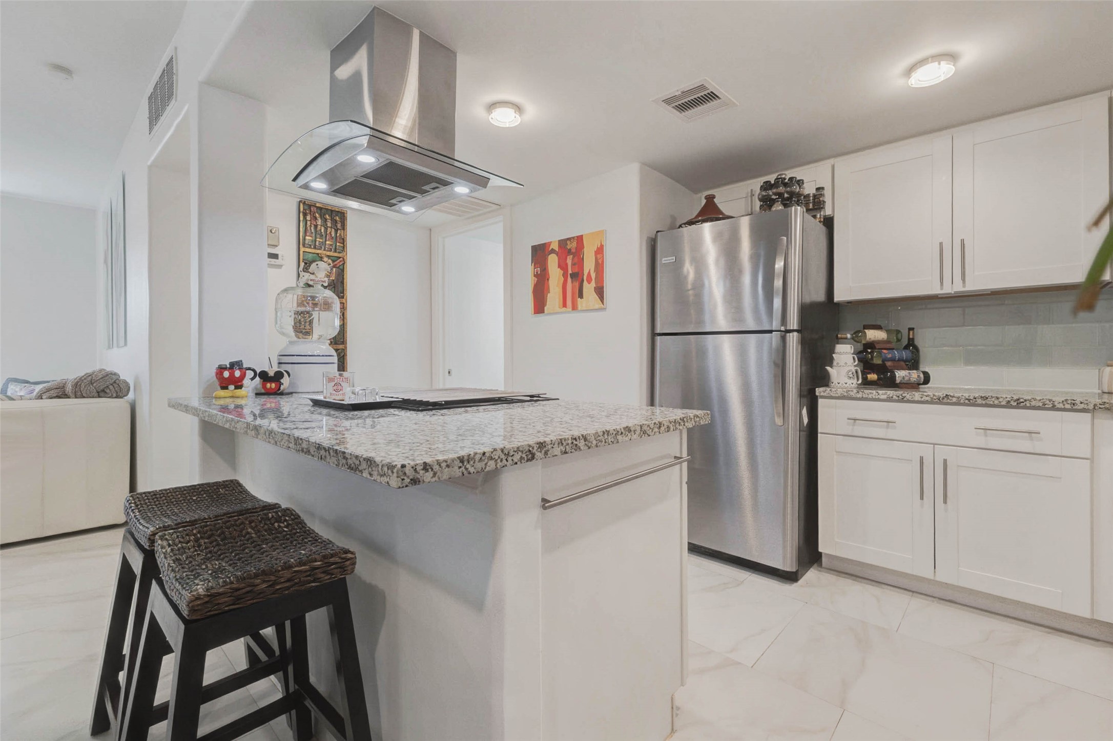 Modern white cabinetry and granite countertops, with a stainless steel refrigerator. It includes a breakfast bar area with seating and an overhead range hood, creating a functional and stylish space.