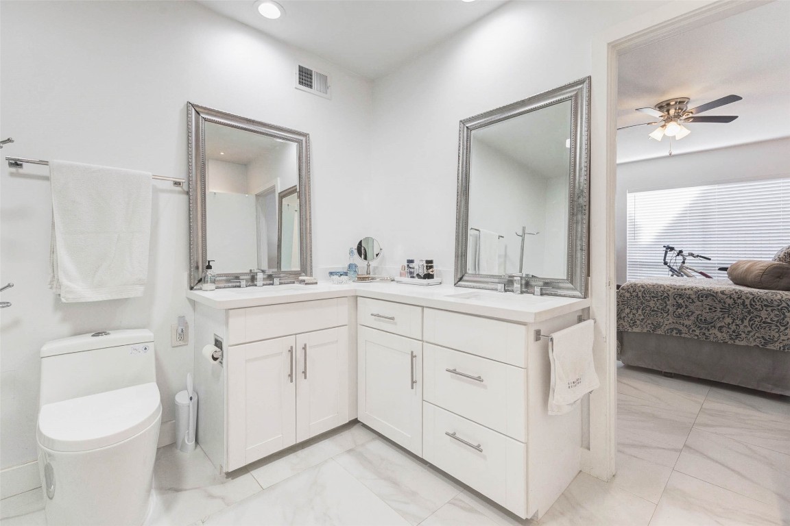 2234 South Piney Point Road, Unit 110 Houston, TX 77063 - Photo 19 of 25 Modern bathroom with white cabinetry and dual sinks, featuring large frameless mirrors. The room is well-lit with recessed lighting, and the design flows seamlessly into a bedroom with a ceiling fan and a large window.