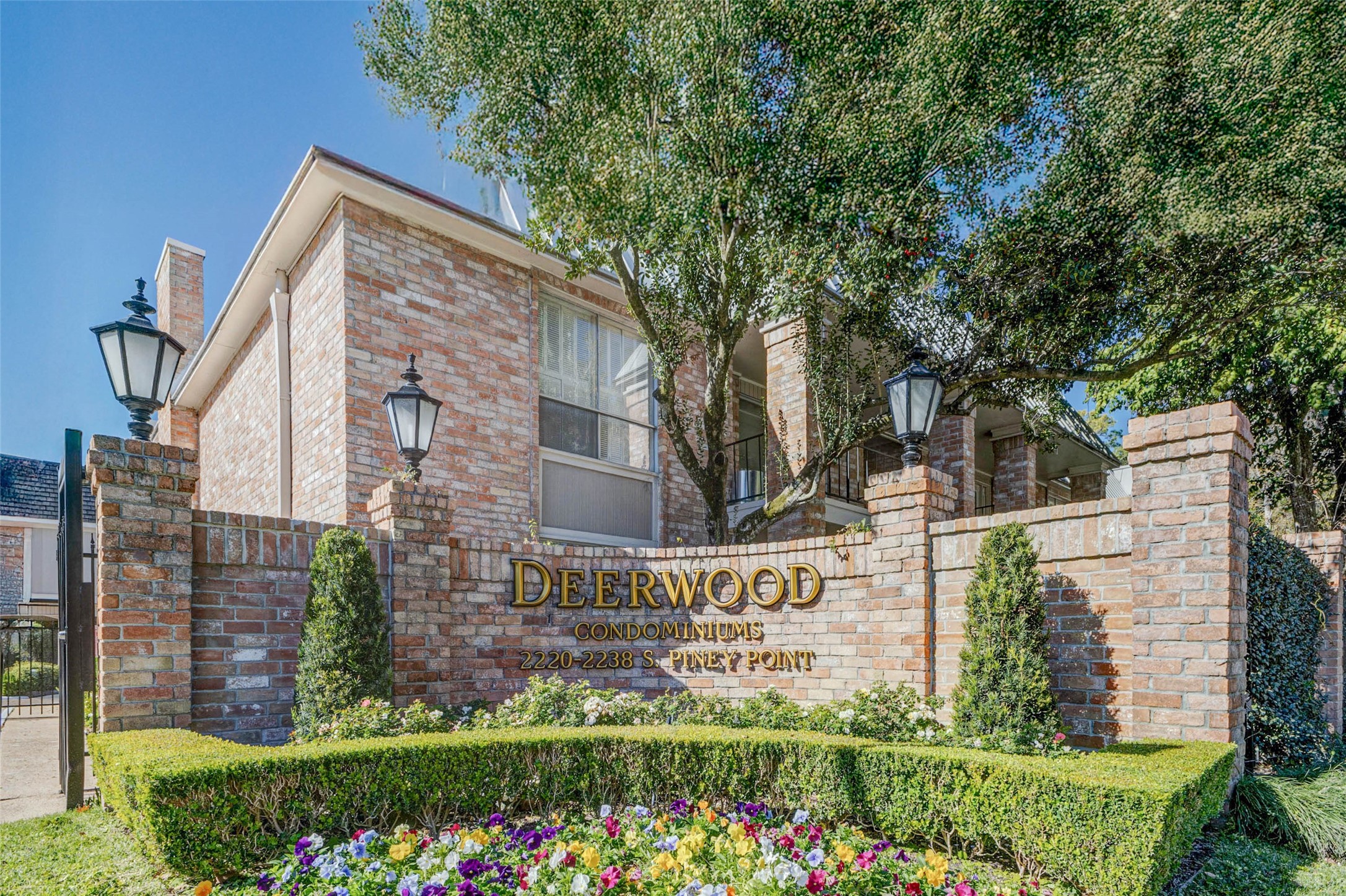 2234 South Piney Point Road, Unit 110 Houston, TX 77063 - Photo 2 of 25 Deerwood Condominiums, featuring a charming brick façade with classic lantern-style lighting. The well-maintained landscaping includes colorful flowers and neatly trimmed hedges, creating an inviting atmosphere.