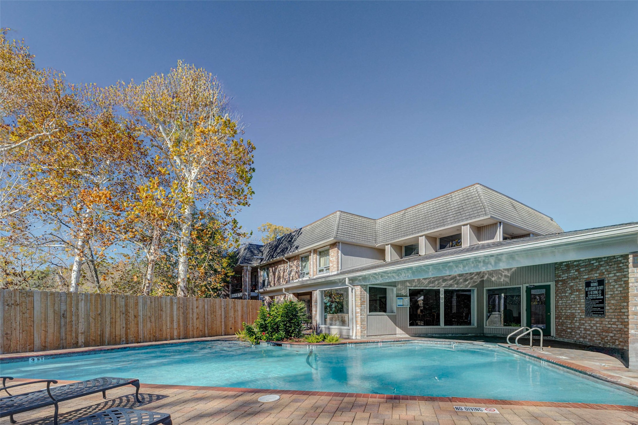 2234 South Piney Point Road, Unit 110 Houston, TX 77063 - Photo 4 of 25 Beautiful pool area adjacent to a clubhouse with large windows. The pool is surrounded by a brick patio with lounge chairs and has a wooden privacy fence. Mature trees provide a natural backdrop, creating a serene environment. The condo is few minutes, you could see it through the balcomy.