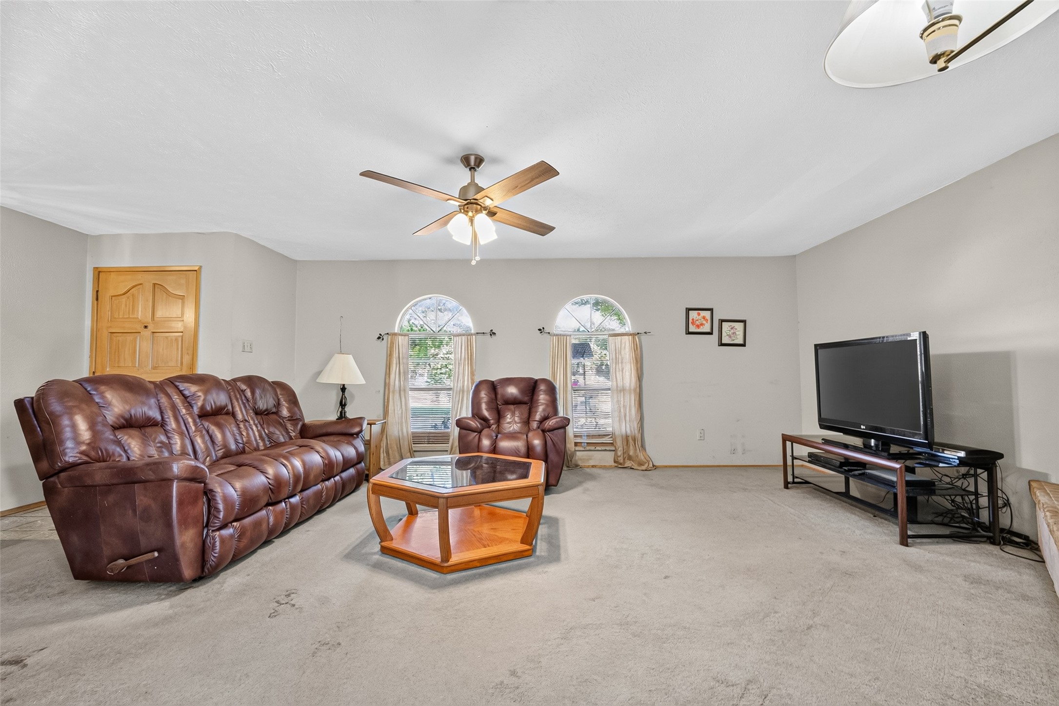 22926 Millgate Drive Spring, TX 77373 - Photo 6 of 34 Spacious living room with neutral tones, featuring a ceiling fan, large windows for natural light, and comfortable leather seating. It includes a modern entertainment setup and a central coffee table, creating a cozy and inviting atmosphere.