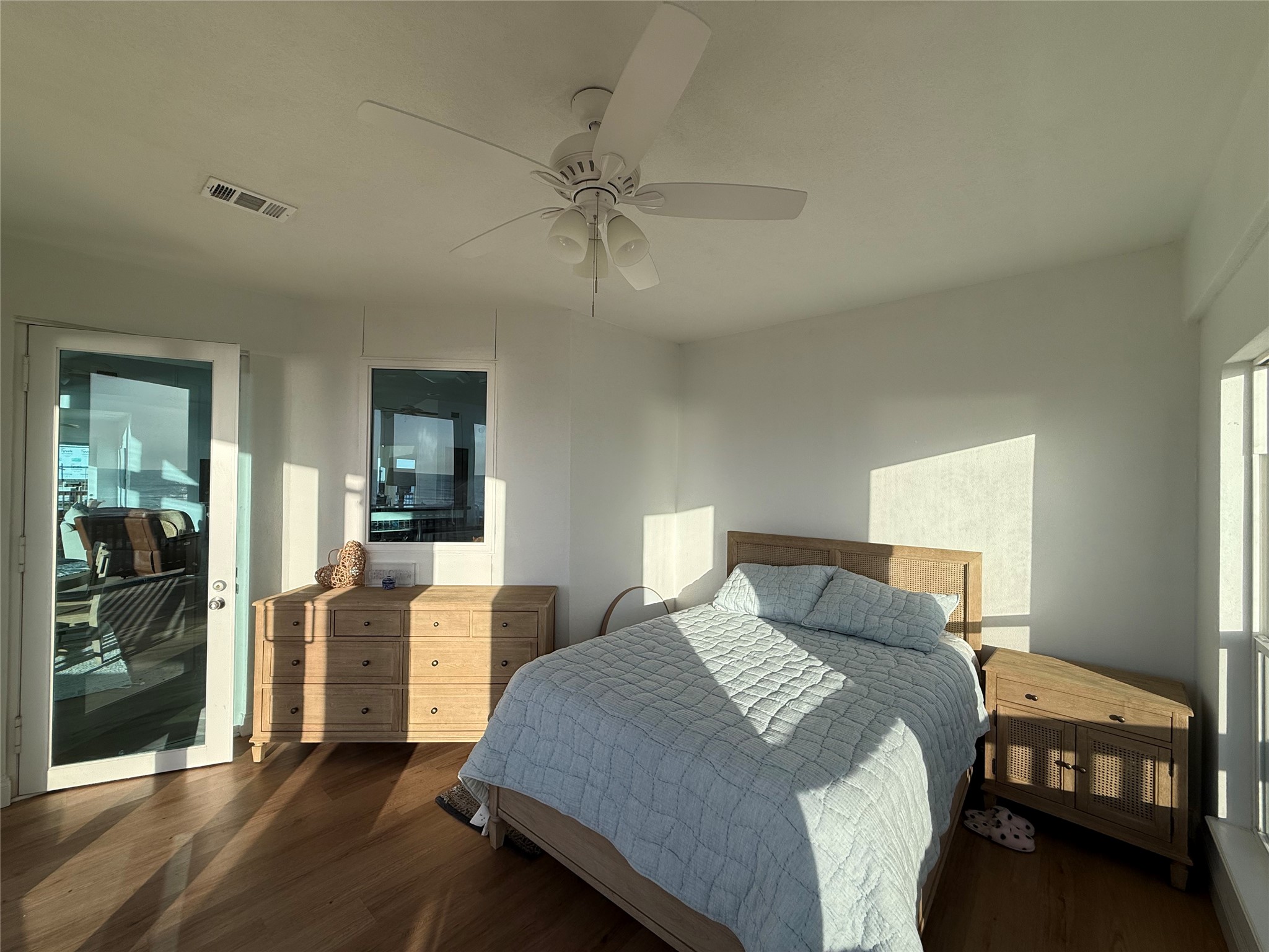 878 County Road 230 Sargent, TX 77414 - Photo 25 of 39 Bright bedroom with natural light, featuring a ceiling fan, hardwood floors, and light wood furniture. There's a cozy bed, dresser, and nightstand, with a door leading to a patio or balcony area.