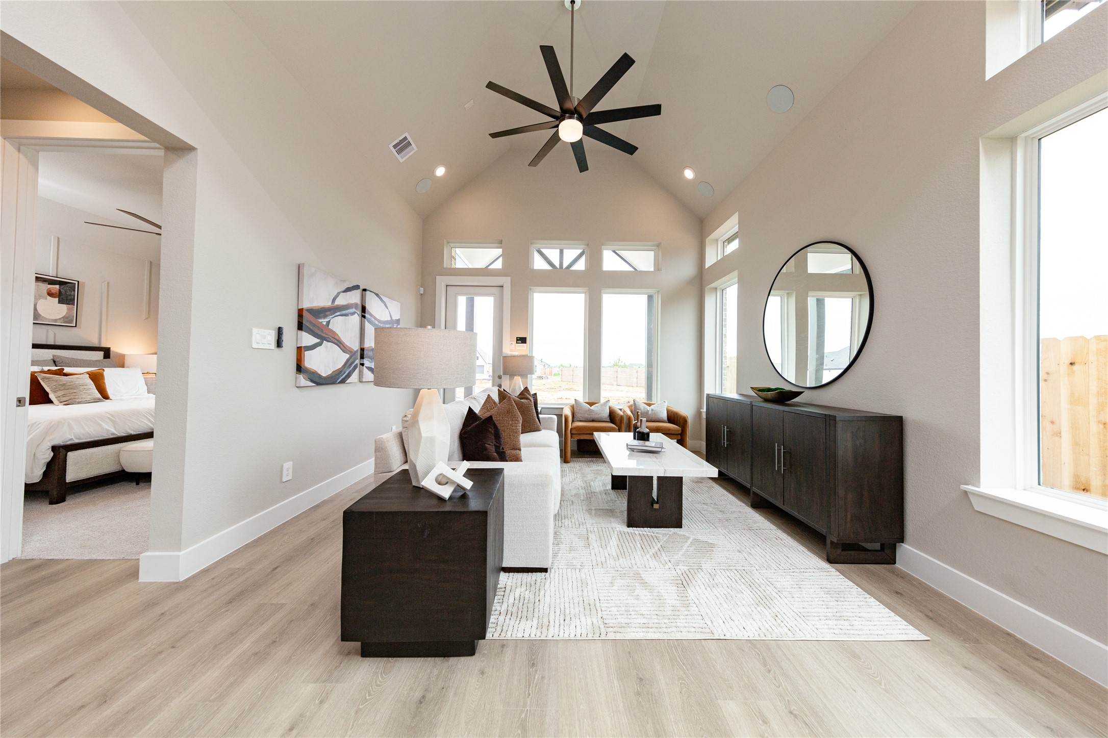 This expansive family room features dramatic vaulted ceilings accented by a large, modern multi-blade ceiling fan that creates an open and majestic feel. A wall of towering windows, topped with beautiful transom windows, drenches the entire space in bright natural light and includes a direct glass exterior door to the backyard area. Wide-plank flooring stretches throughout the level, seamlessly connecting the grand living area to the adjacent primary bedroom suite.