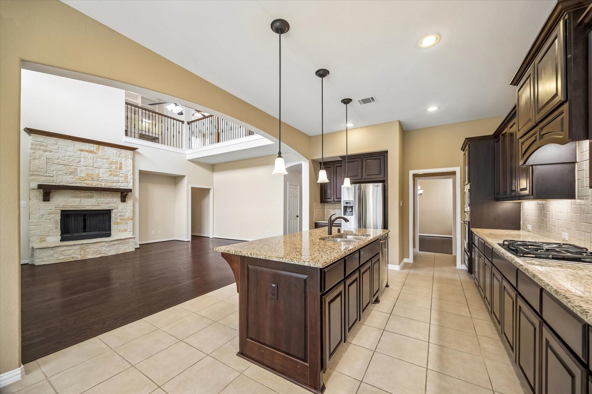 17403 Astrachan Road Richmond, TX 77407 - Photo 9 of 40 This spacious open-concept kitchen and living area features a large island with granite countertops, dark wood cabinetry, and modern appliances. The adjacent living space has a stone fireplace and high ceilings with a view of the upstairs balcony, offering a warm and inviting atmosphere.