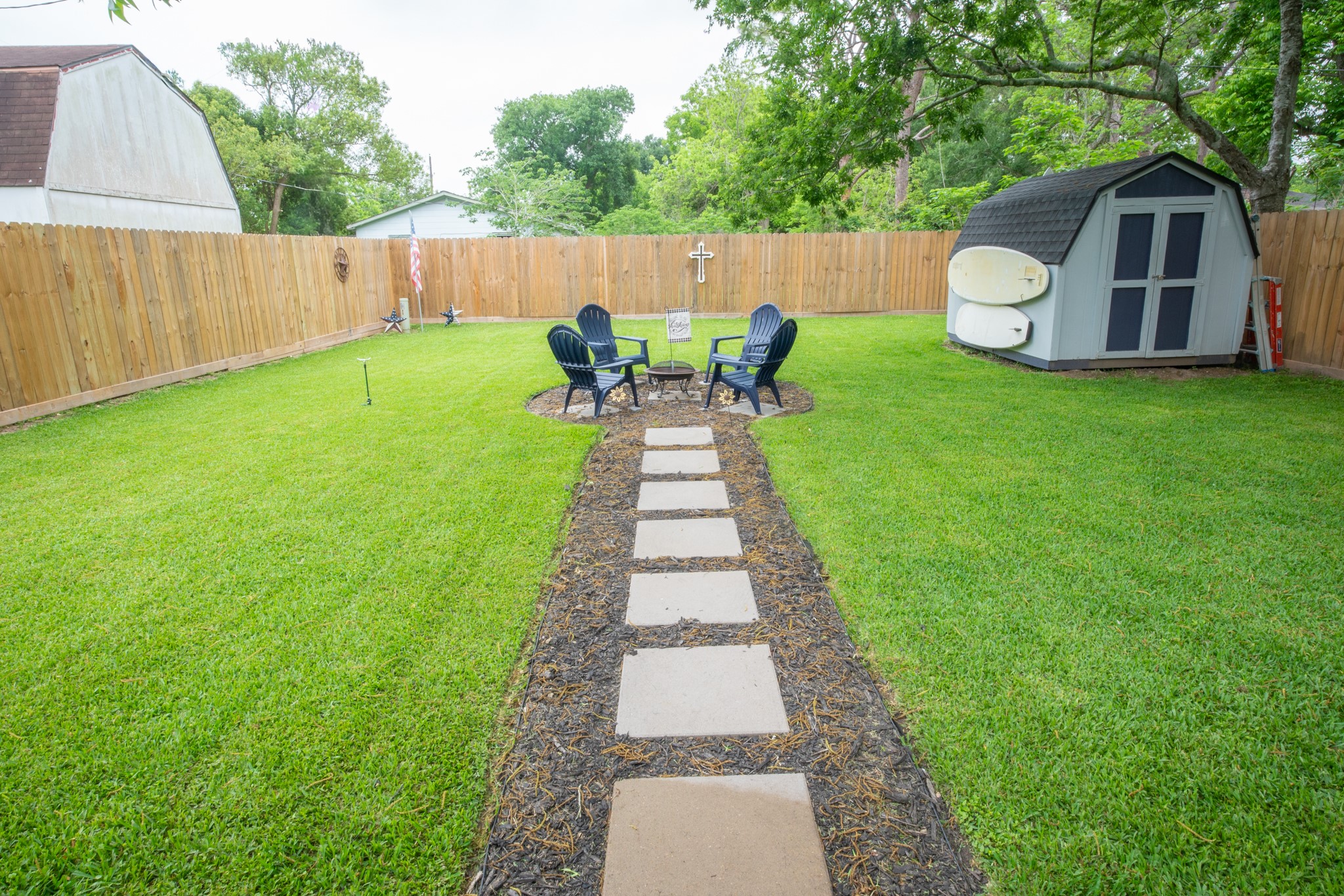 846 South Anderson Street Angleton, TX 77515 - Photo 3 of 30 A well-maintained backyard featuring a grassy lawn, a stone path leading to a seating area with chairs around a fire pit, and a small storage shed. Perfect for outdoor relaxation and gatherings.