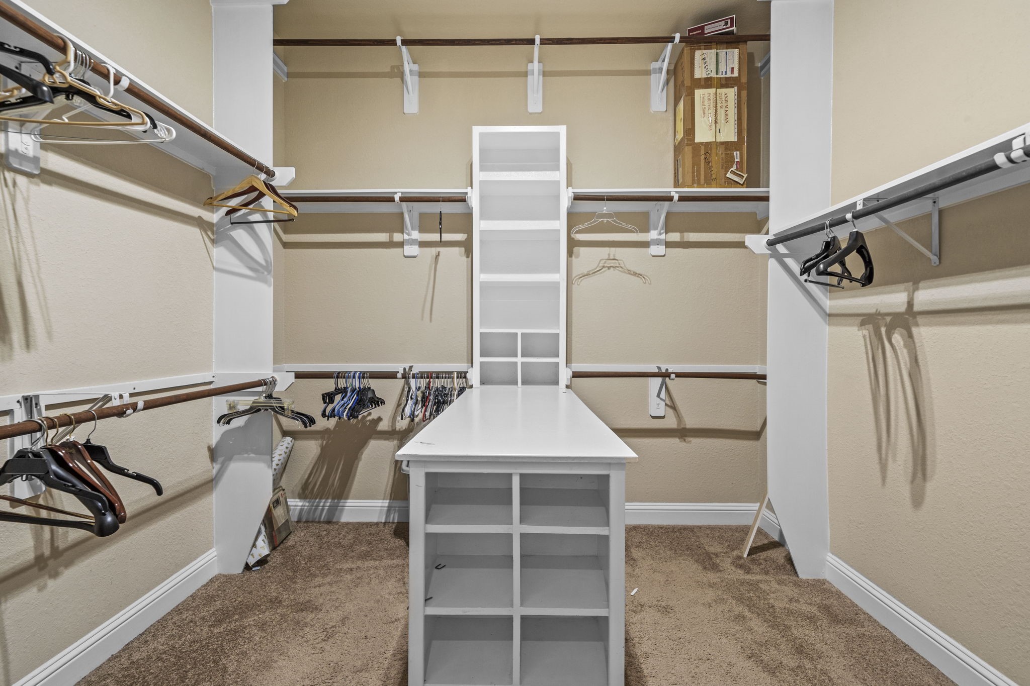 14215 Spindle Arbor Road Cypress, TX 77429 - Photo 34 of 47 This photo shows a spacious walk-in closet with beige walls and carpeted flooring. It features multiple wooden rods for hanging clothes, several shelves for storage, and a central white shelving unit with additional compartments. The space is well-organized and offers ample storage.