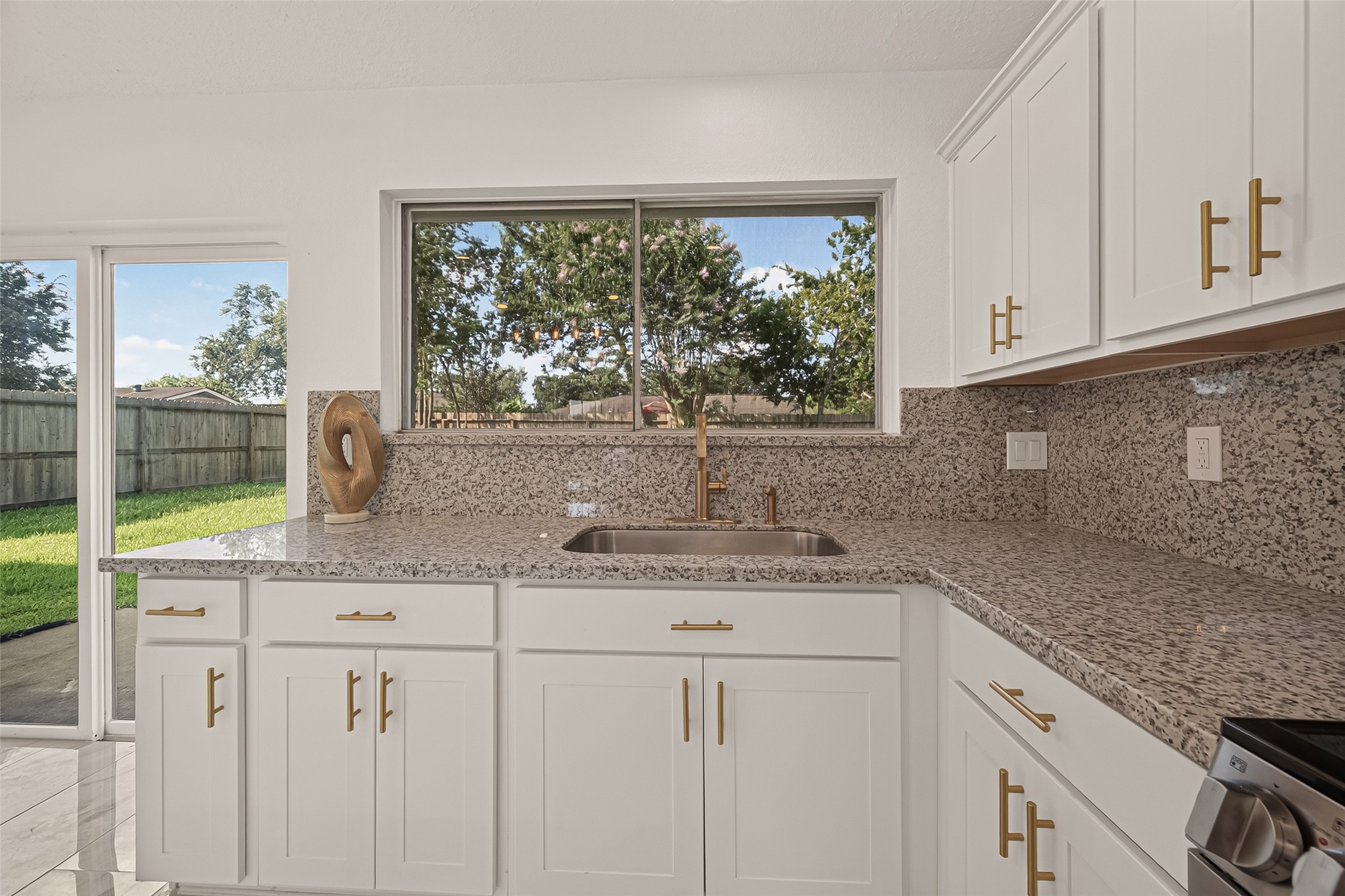 16619 Tibet Road Friendswood, TX 77546 - Photo 8 of 29 This kitchen features elegant granite countertops, white cabinetry with gold hardware, and a large window offering a view of the backyard. Natural light fills the space, creating a bright and inviting atmosphere.