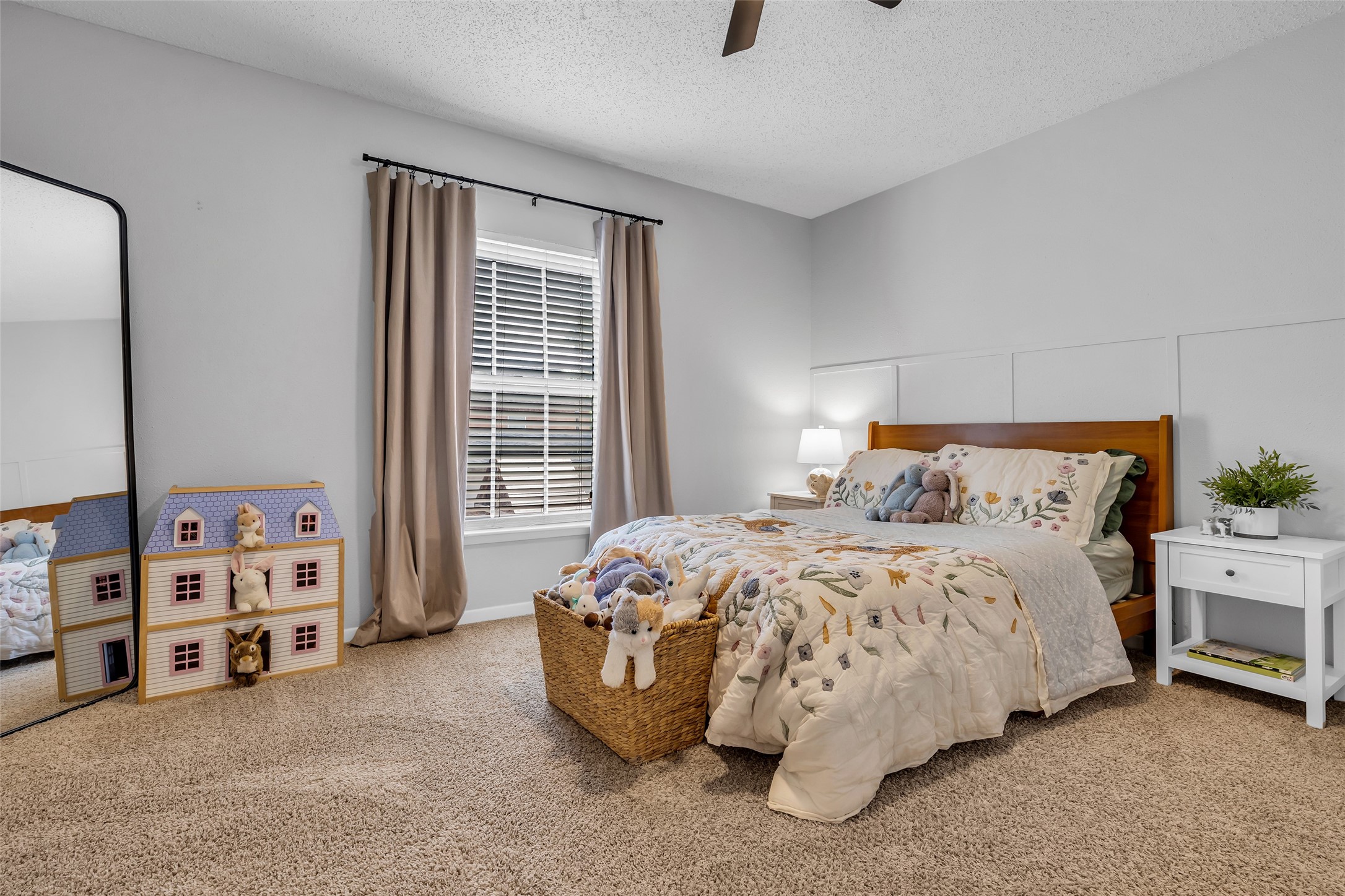 9415 Towerstone Drive Spring, TX 77379 - Photo 33 of 44 This cozy bedroom features a comfortable bed with floral bedding, a bedside table with a lamp, and a large window with curtains for natural light. The carpeted floor adds warmth, and a charming dollhouse sits next to a large mirror, creating a playful yet serene atmosphere.