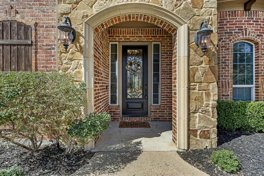 6705 Sigma Lane Frisco, TX 75035 - Photo 2 of 8 Arched stone entryway featuring a dark wood-finish door with decorative glass, flanked by sidelight windows and exterior sconces, set against a brick and stone facade